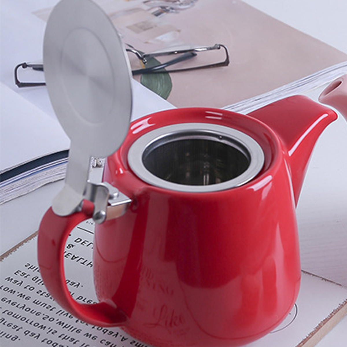Eine rote Teekanne mit silbernem Deckel steht auf einem Tisch. Der Deckel ist offen. Im Inneren befindet sich ein Metallsieb. Im Hintergrund sind Gläser und Papier.