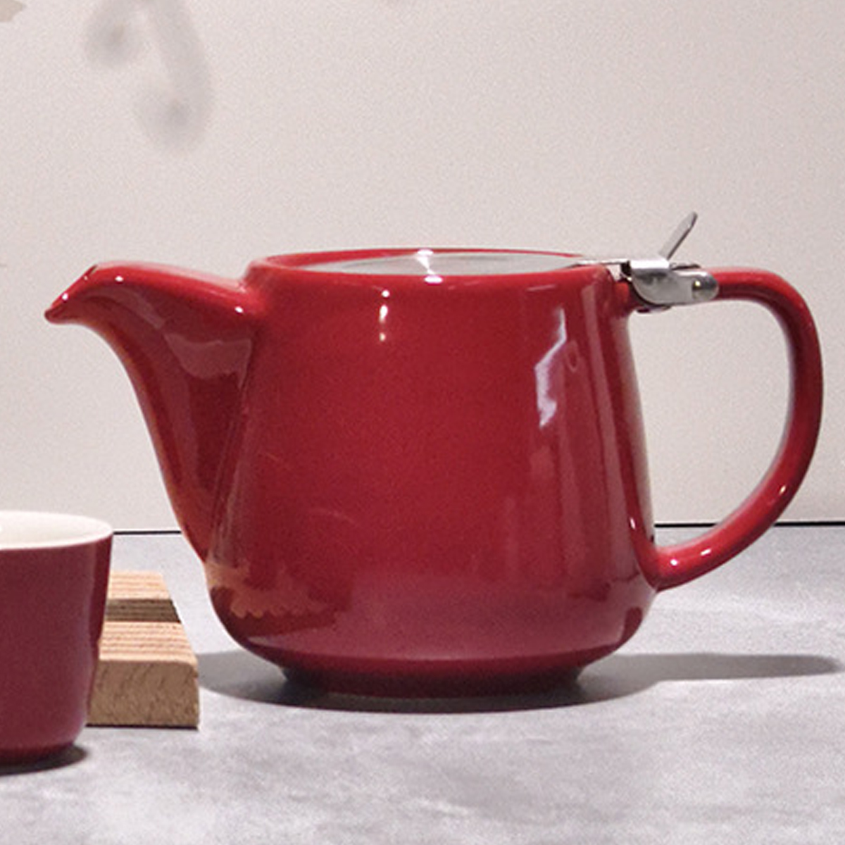 Eine rote Teekanne mit silbernem Griff und Ausguss steht auf einer grauen Oberfläche. Eine Tasse und ein Holzuntersetzer stehen daneben.
