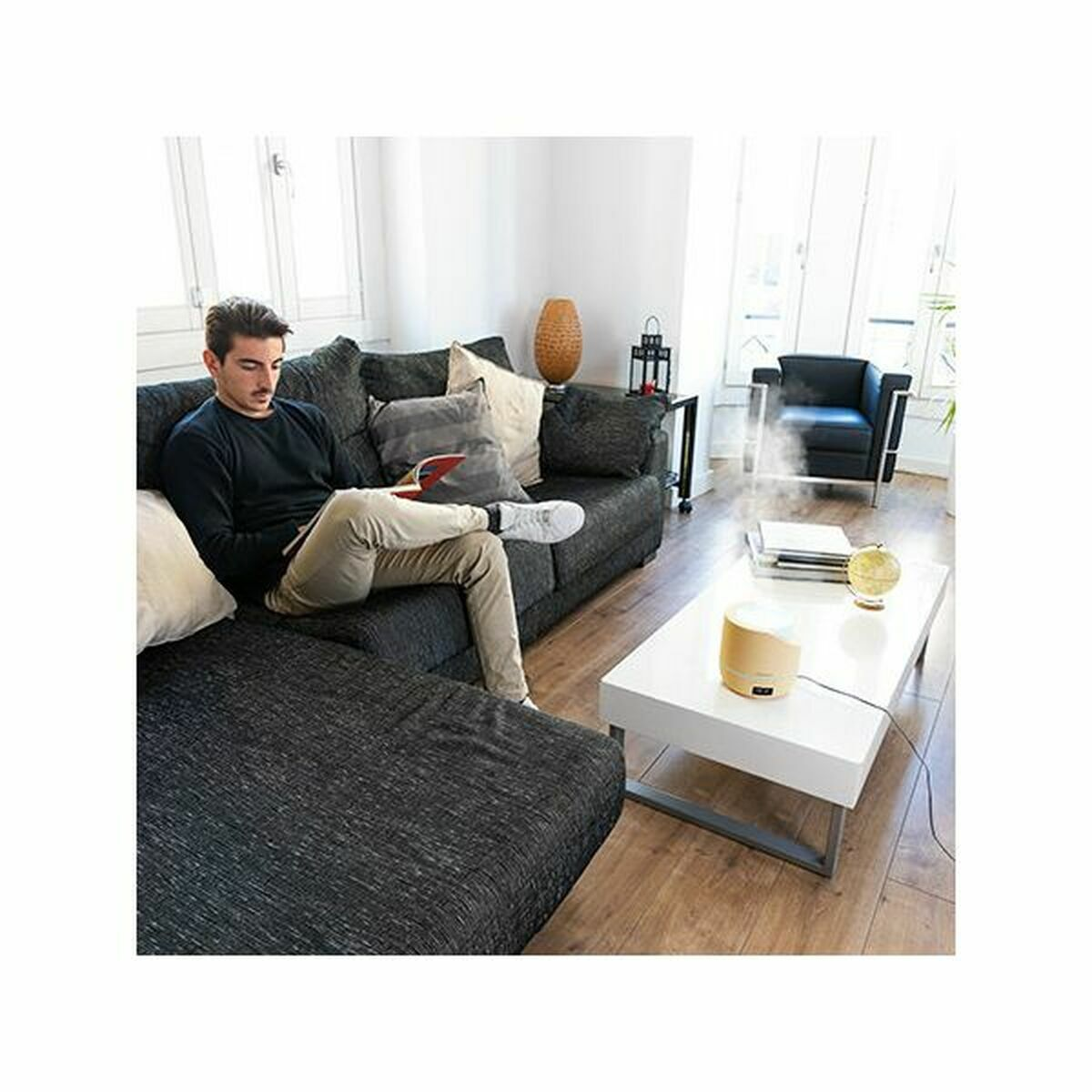 Mężczyzna czyta na kanapie, nawilżacz na białym stole, czarny fotel i dekoracyjny wazon w pokoju.