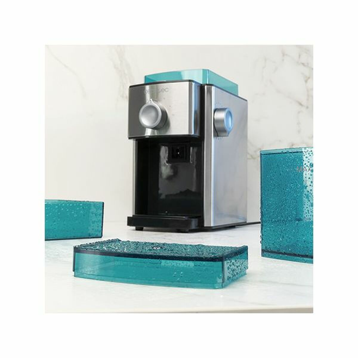 Srebrno-turkusowa maszyna do lodu z trzema turkusowymi tackami na lód na białym blacie. Tło to marmurowa ściana.