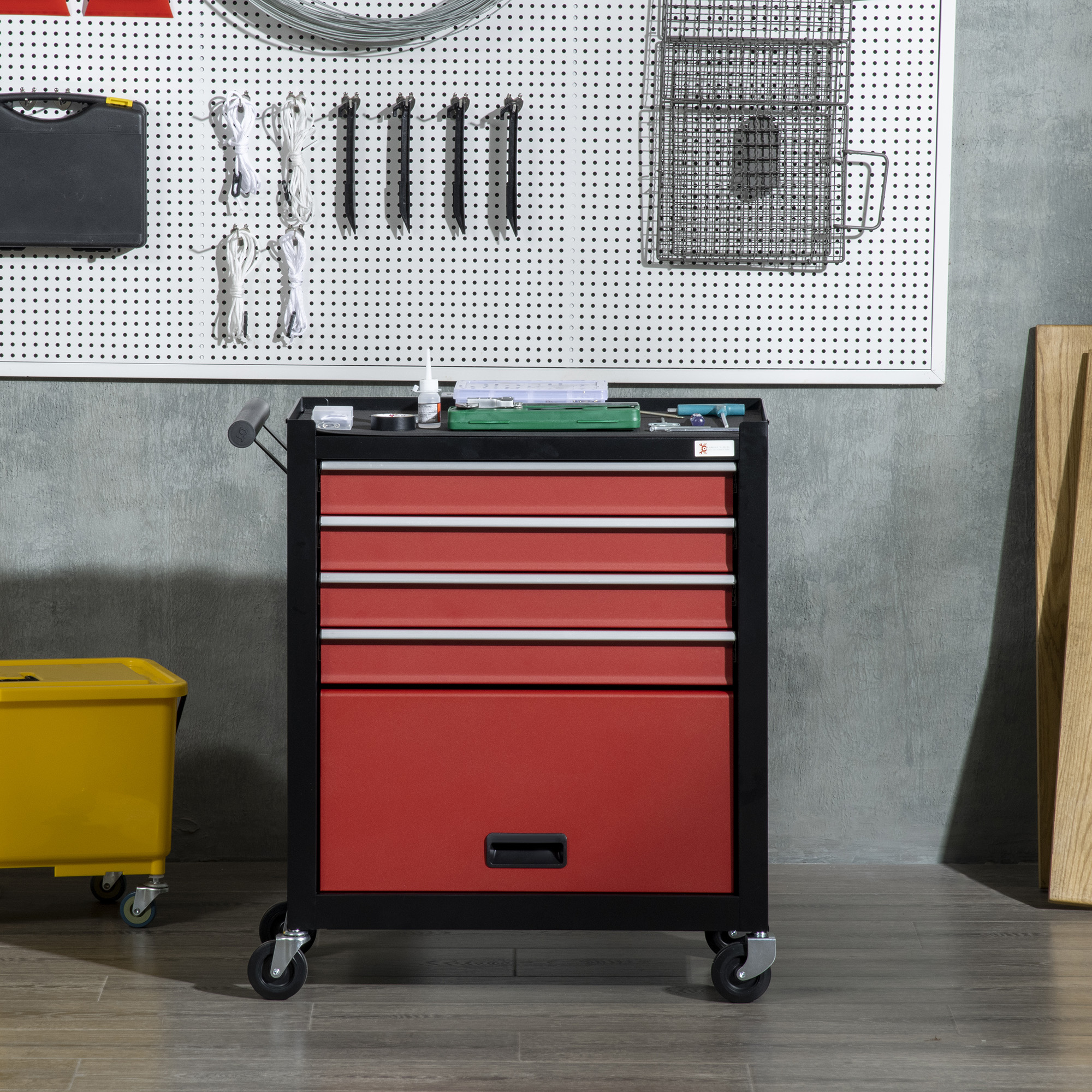 Caja de herramientas roja sobre ruedas, marco negro, con cajones. Herramientas, panel perforado, cubo amarillo y tabla de madera al fondo.