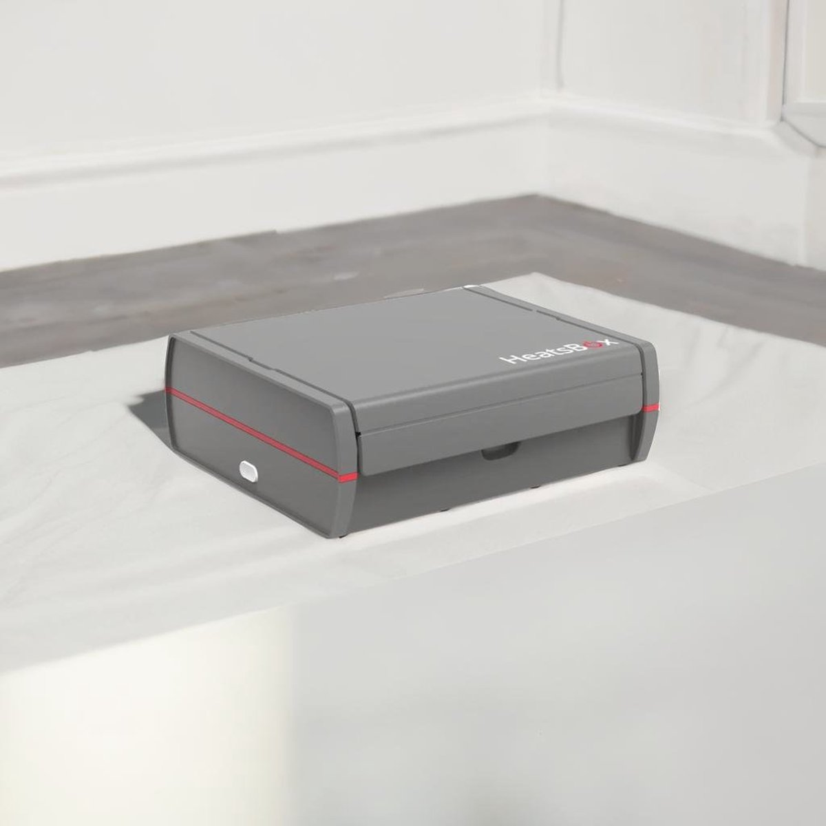 Eine graue HeatsBox liegt auf weißem Stoff. Rote Linien akzentuieren die Box. Der Hintergrund ist eine weiße Wand und ein grauer Boden.