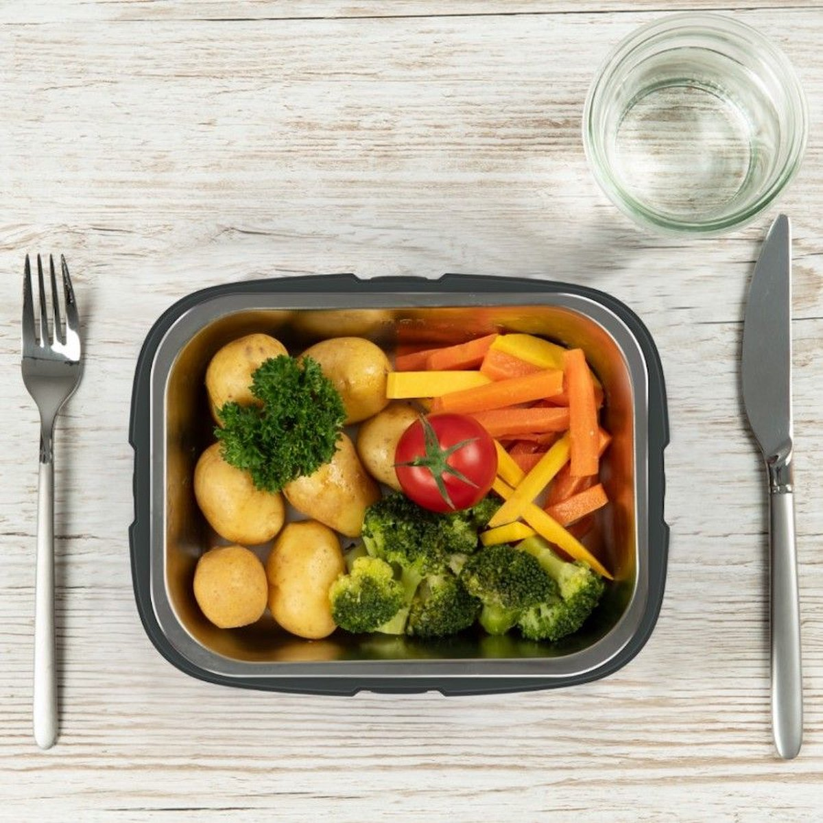 Eine Lunchbox mit Kartoffeln, Karotten, Brokkoli und einer Tomate. Eine Gabel, ein Messer und ein Glas Wasser sind ebenfalls vorhanden.