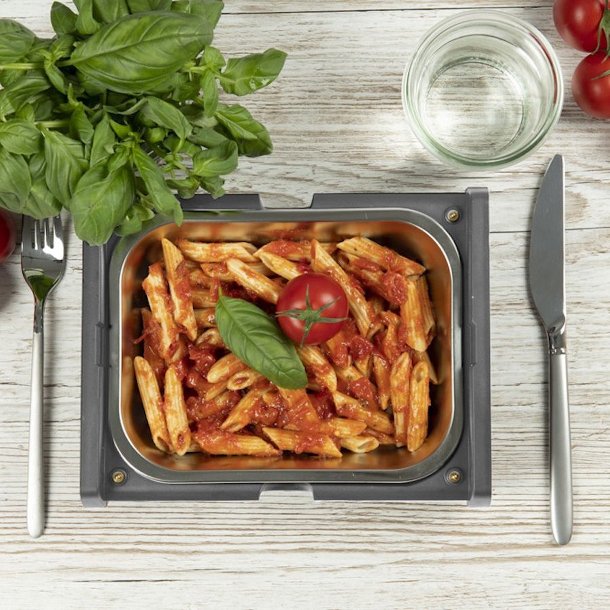 Penne-Pasta mit Tomatensauce, Basilikum und Tomate in einem Metallbehälter, mit Messer, Gabel und Wasserglas auf einem Holztisch.