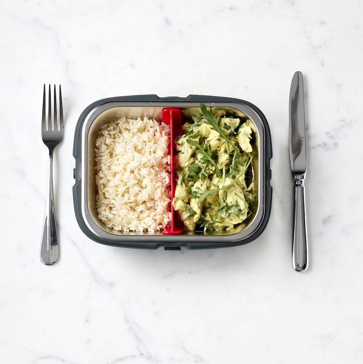 Geteilte Lunchbox mit Reis und Hühnchen. Gabel und Messer liegen auf weißer Marmoroberfläche.