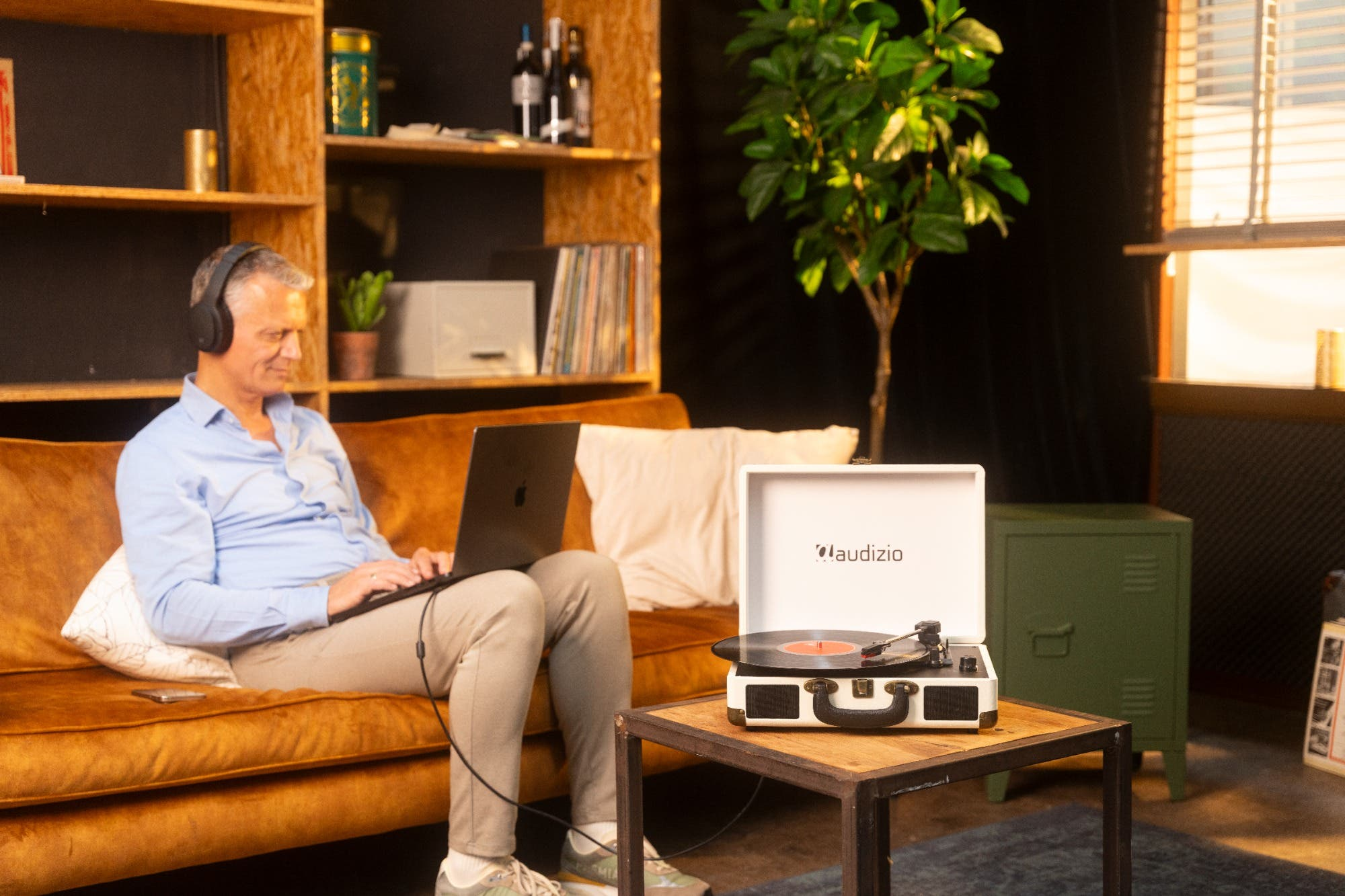 Hombre con auriculares en un sofá usando una laptop. Un tocadiscos está sobre una mesa. Estanterías y una planta en el fondo.