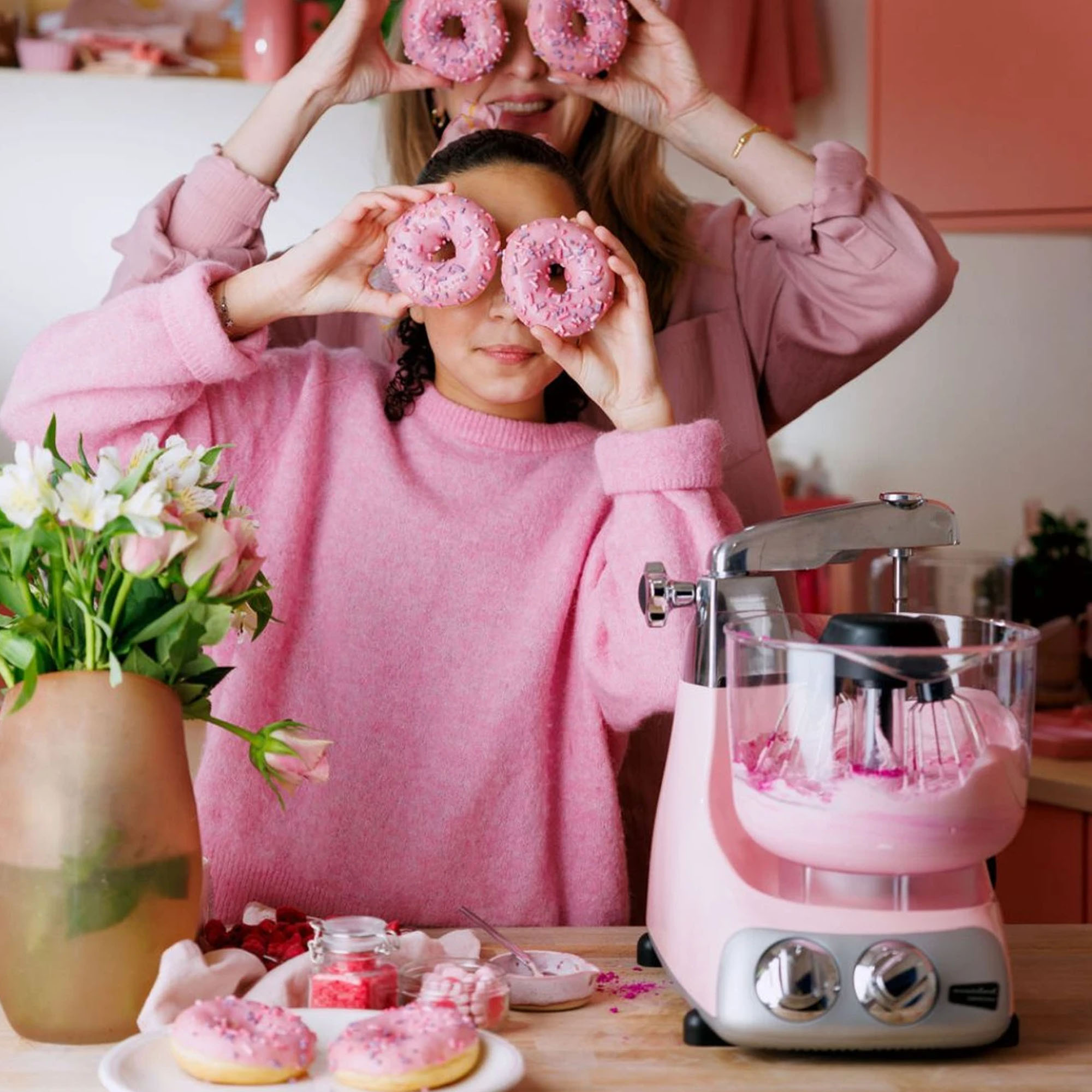 İki kadın gözlerinin üzerine donut tutuyor. Pembe bir mikser, çiçekler ve daha fazla donut masada.