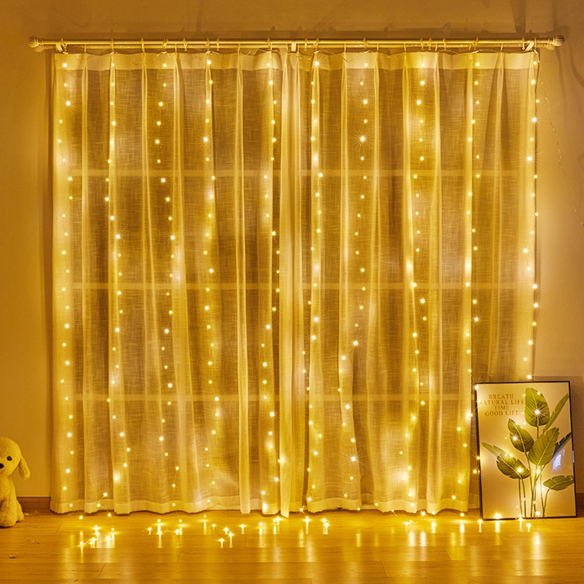 Ein Vorhang aus Lichterketten hängt vor einem Fenster. Ein gerahmtes Bild und ein Stofftier befinden sich rechts und links.