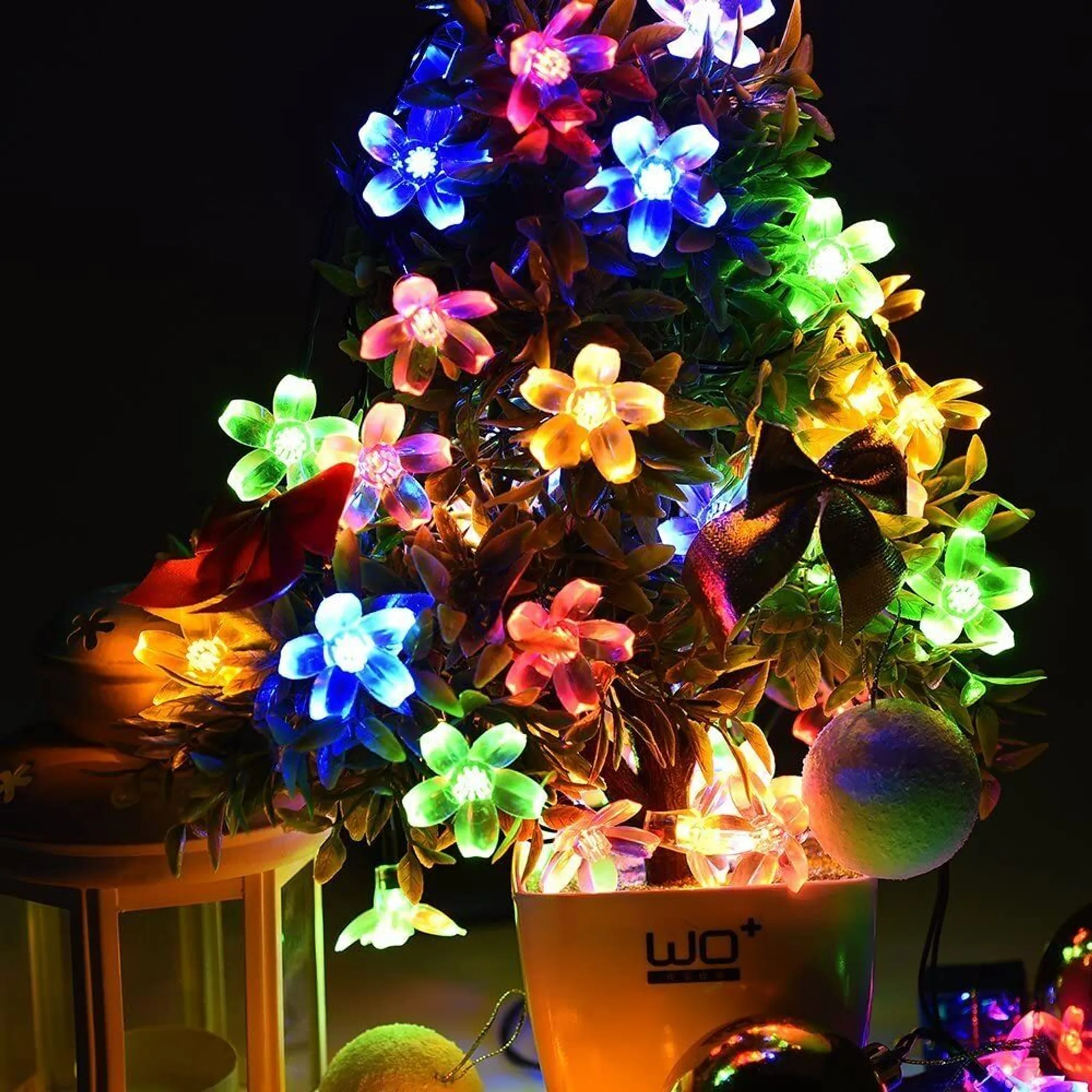 Ein kleiner Weihnachtsbaum mit blütenförmigen Lichtern in verschiedenen Farben. Eine Schleife und Ornamente sind ebenfalls sichtbar.