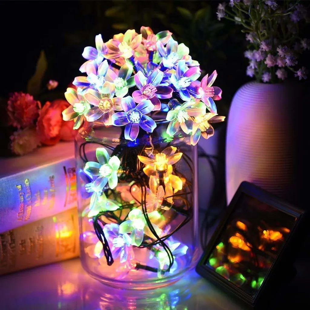 Ein Glas mit blütenförmigen Lichtern. Weitere Objekte sind ein Buch, Blumen, eine Vase und ein kleiner Spiegel. Alles ist beleuchtet.
