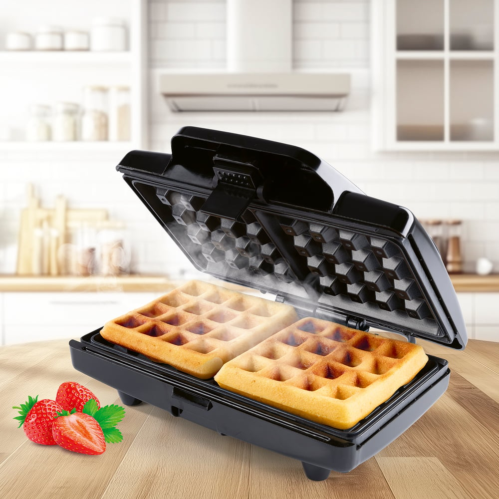 Ein Waffeleisen mit zwei Waffeln, Dampf steigt auf. Erdbeeren und ein Holztisch im Vordergrund. Küchenhintergrund.