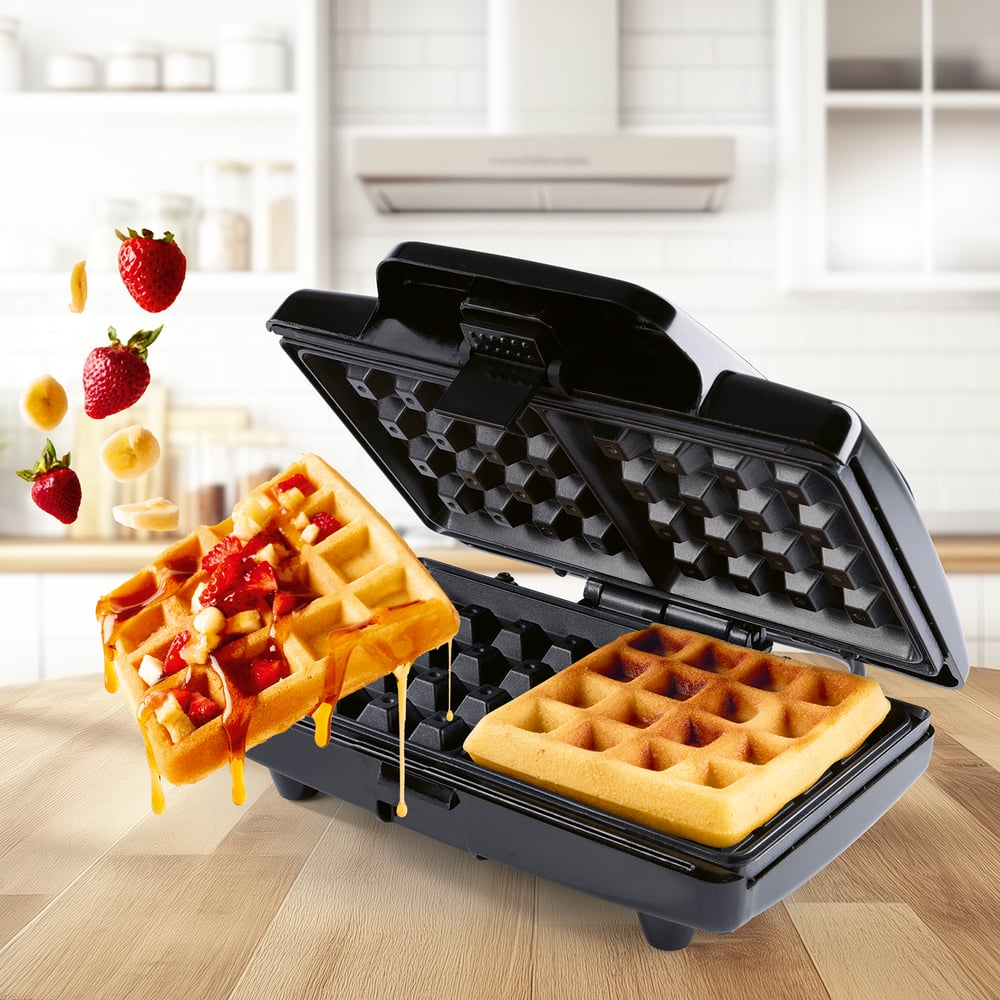 Waffeleisen mit Waffeln, Sirup und Früchten. Waffeln sind im Eisen und schweben mit Erdbeeren und Bananen.