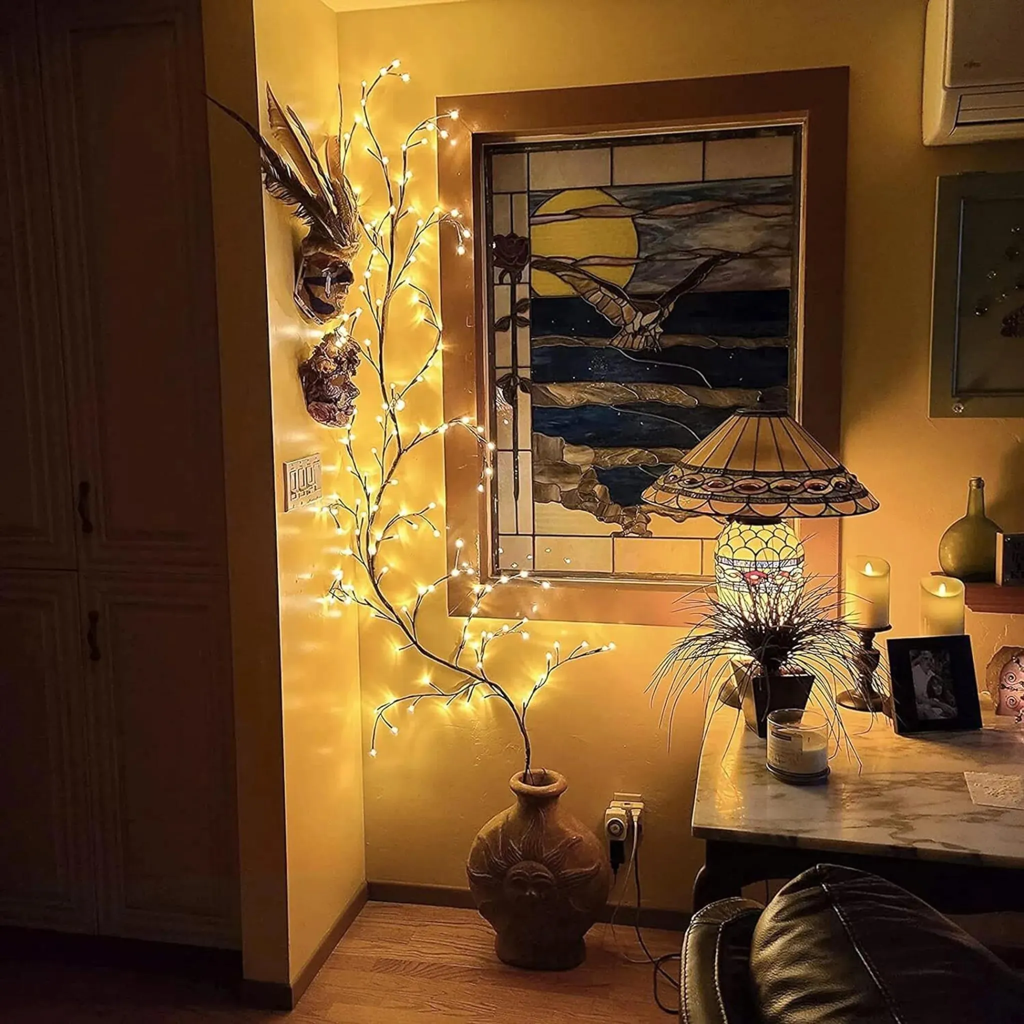 Warmes Zimmer mit beleuchteten Ästen, Buntglasfenster, Lampe, Tisch und Kerzen. Weiche Beleuchtung schafft eine gemütliche Atmosphäre.