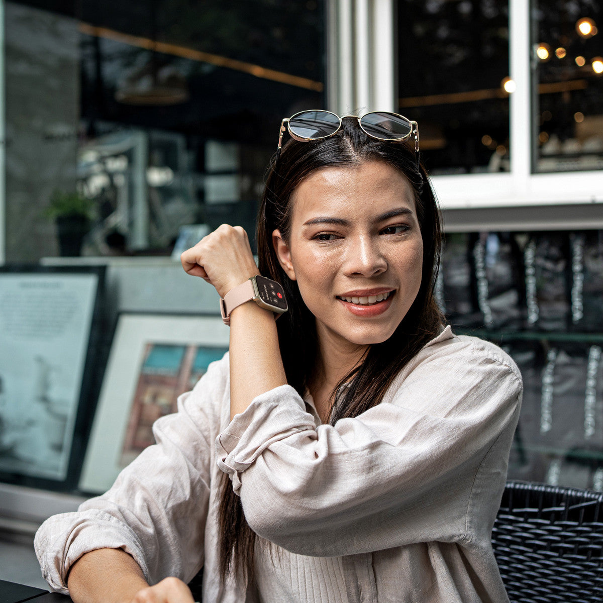 Una donna con smartwatch sorride mentre guarda l'orologio. Occhiali da sole sulla testa. Sfondo: un bar.