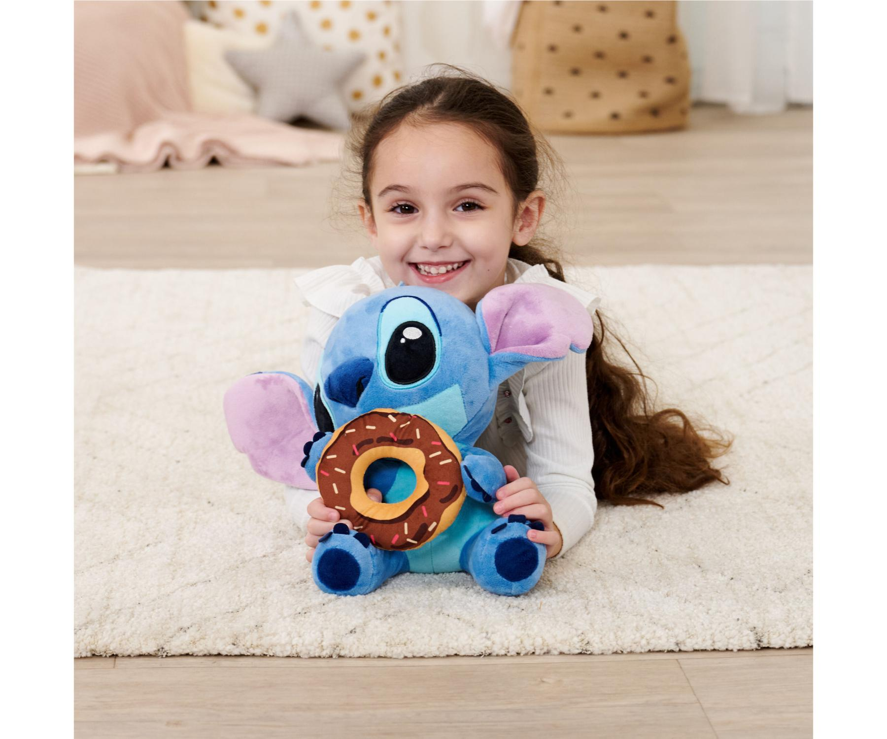 Ein lächelndes Mädchen hält ein Stitch-Plüschtier mit einem Donut. Das Plüschtier liegt auf einem weißen Teppich, im Hintergrund Kissen und ein Korb.