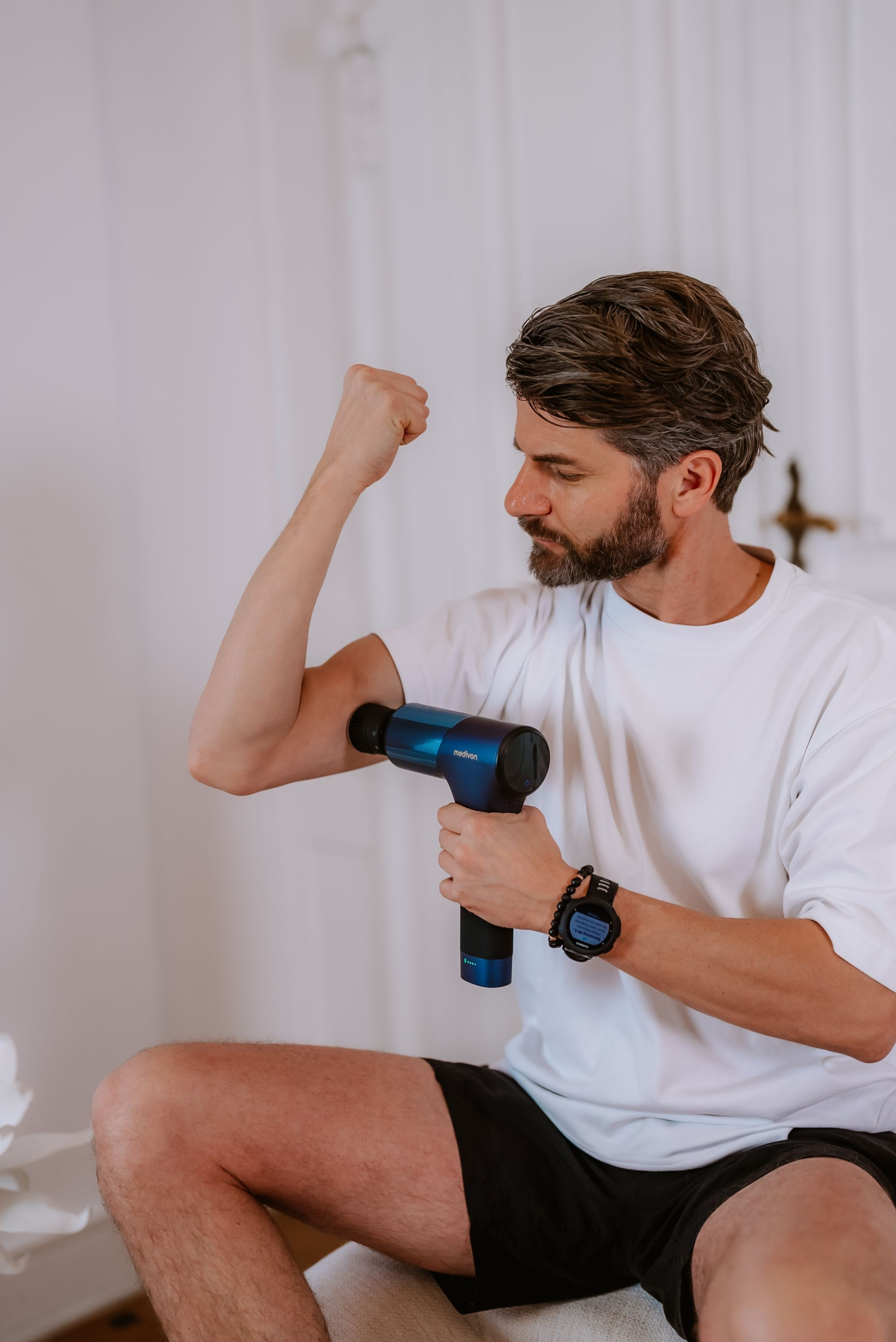 Homme utilisant un pistolet de massage sur son bras. Il porte un t-shirt blanc, un short noir et une montre. Mur blanc en arrière-plan.