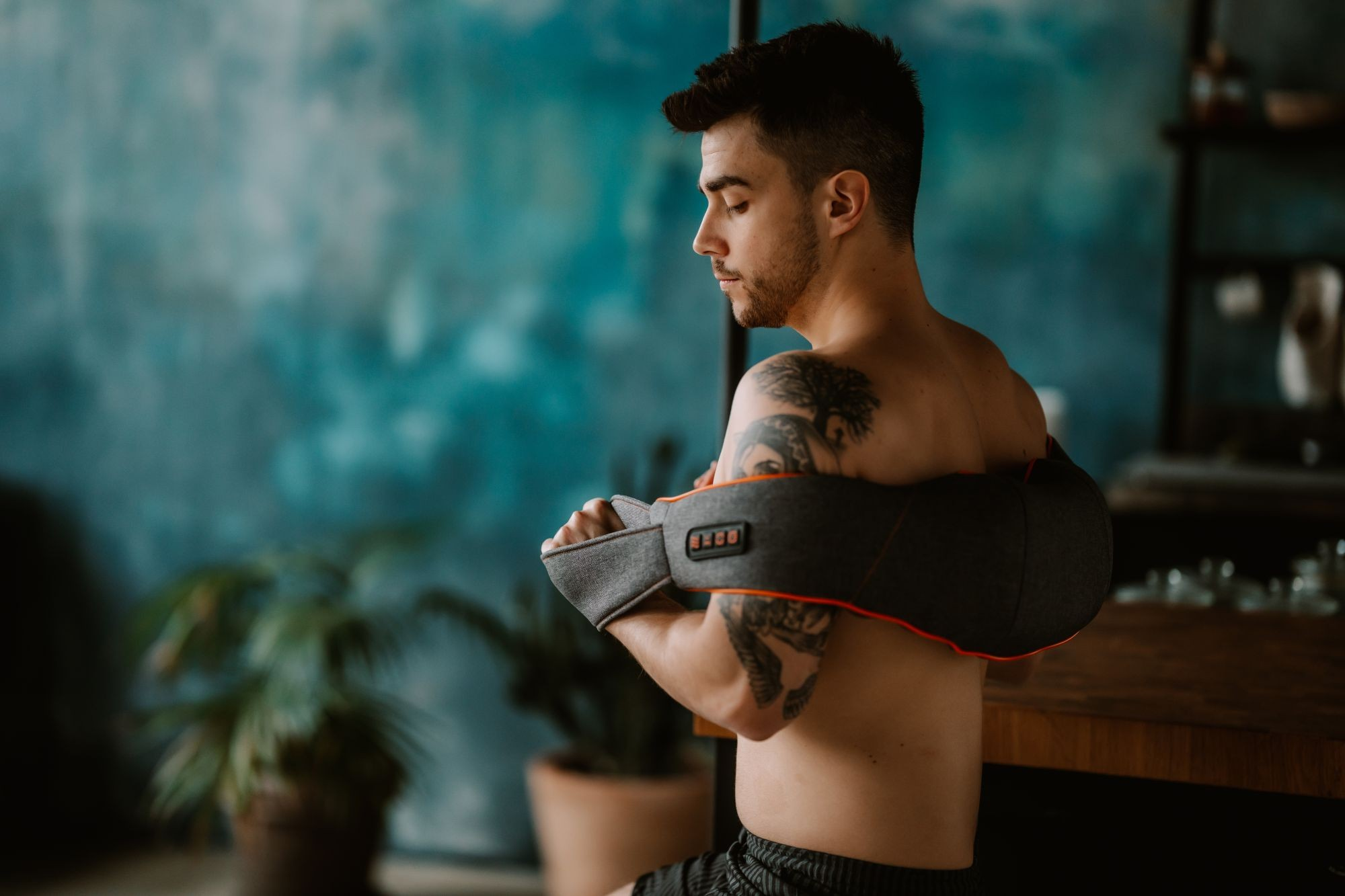 Hombre usando un masajeador de hombros. Tiene tatuajes. El fondo tiene una pared azul y una planta.