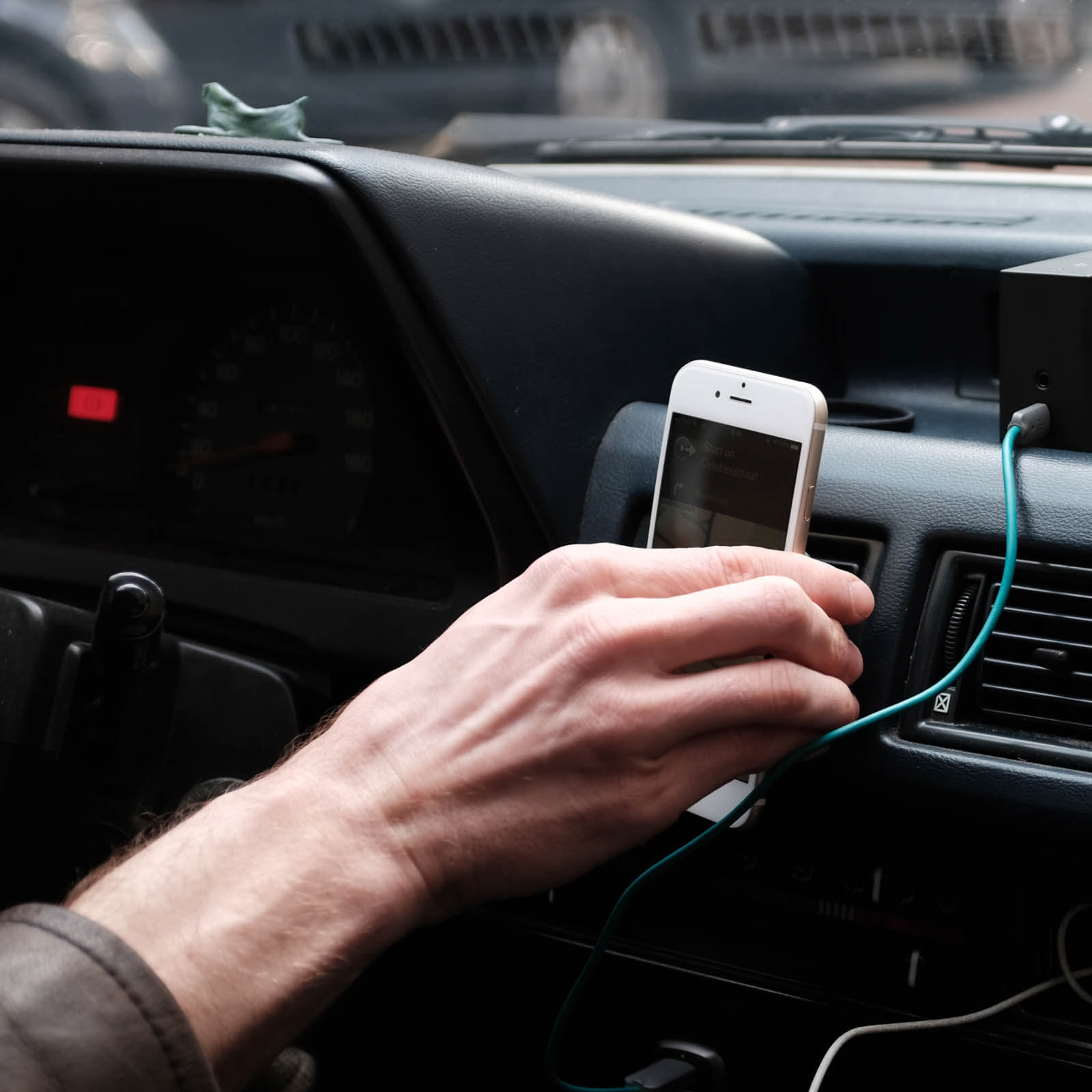 Una mano tiene in mano un telefono bianco in un'auto. Un cavo verde collega il telefono a una scatola nera. Il cruscotto è visibile.