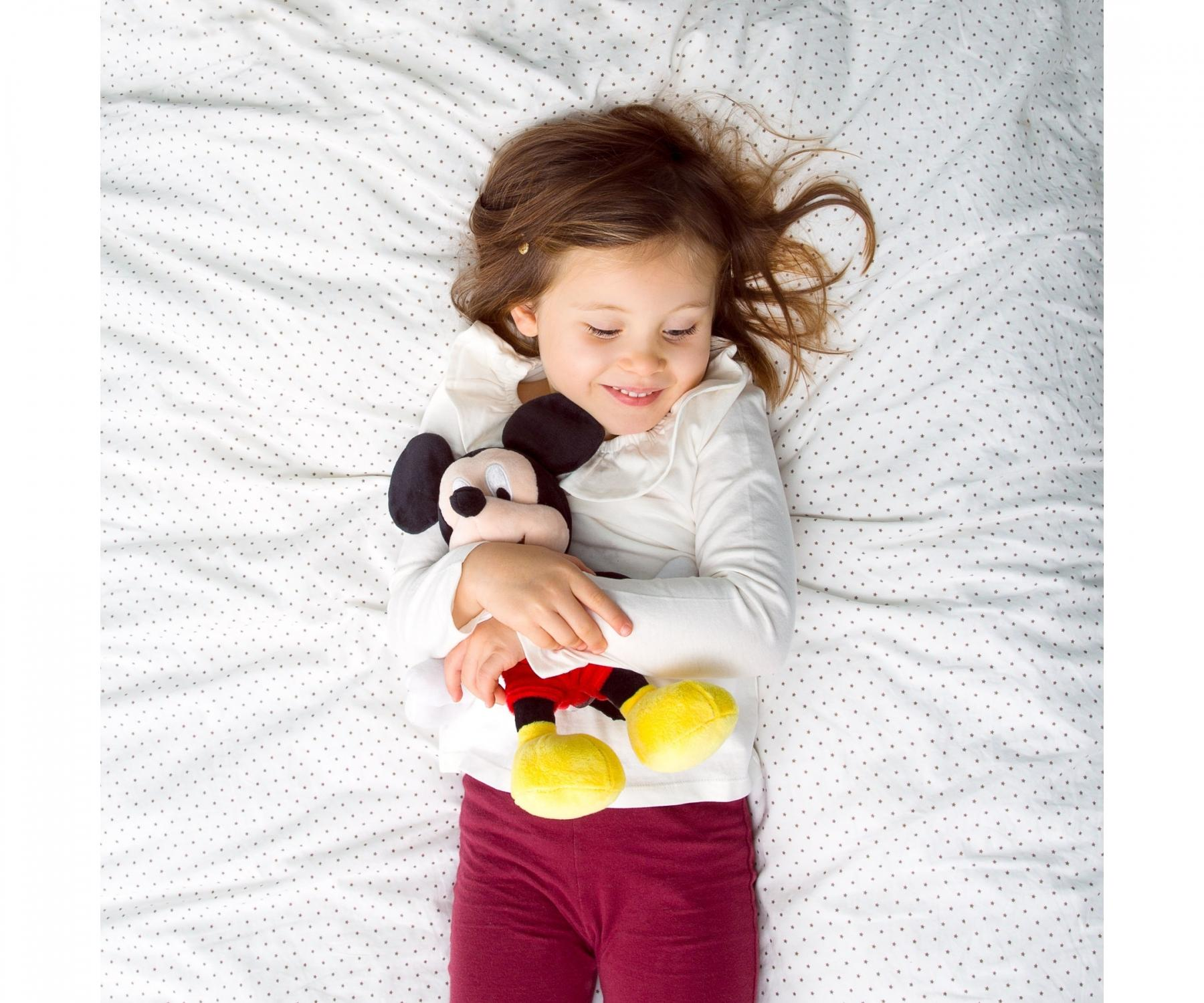 Ein Mädchen liegt auf einem Bett und umarmt ein Mickey-Mouse-Plüschtier. Sie trägt ein weißes Hemd und rote Hose. Das Bettlaken hat ein gepunktetes Muster.