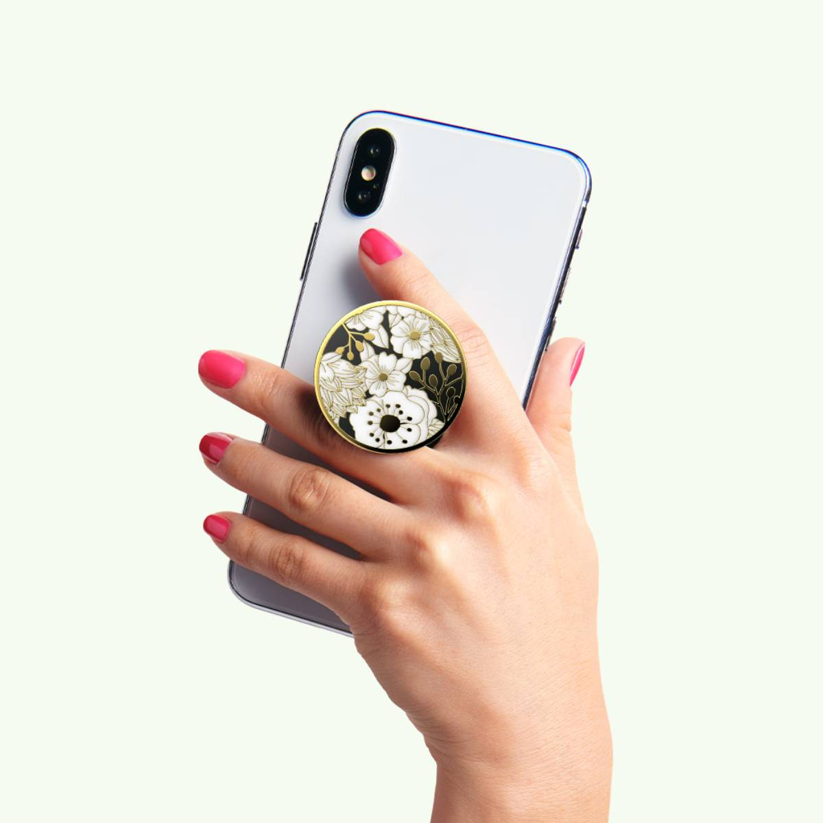 Una mano tiene un telefono bianco con un pop socket floreale. Il pop socket ha fiori bianchi, dettagli dorati e sfondo nero.