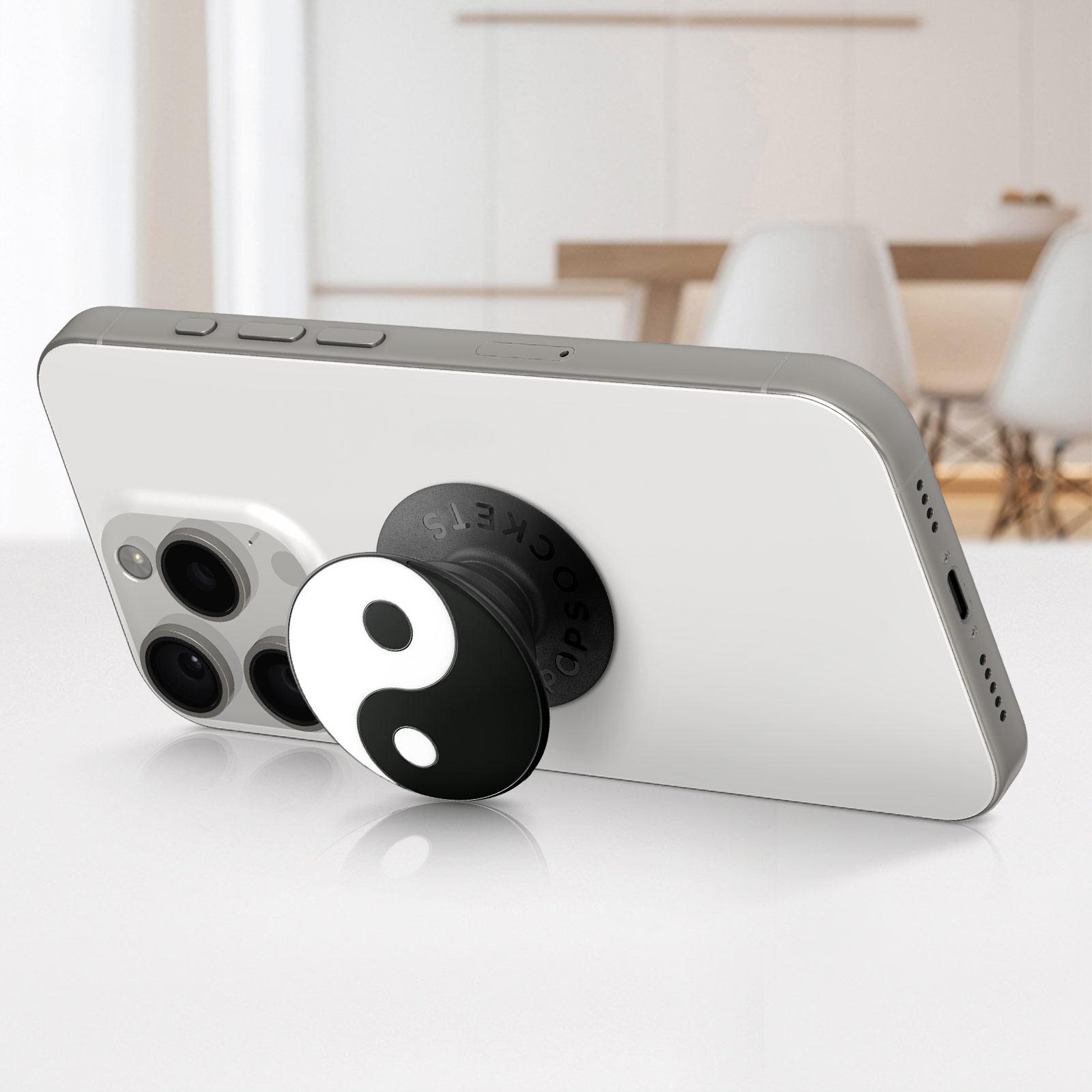 Un teléfono blanco con un PopSocket Yin-Yang. El teléfono está sobre una mesa. El fondo muestra una mesa y sillas.