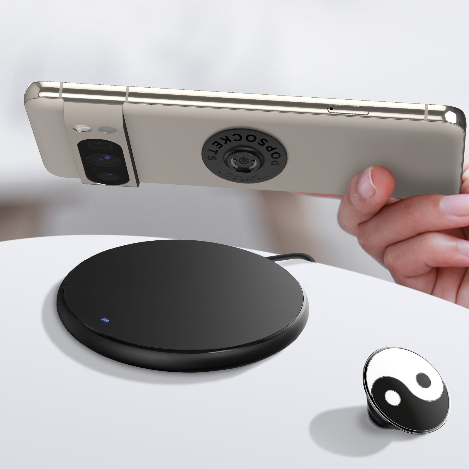Un teléfono con un PopSocket se sostiene sobre un cargador inalámbrico y un PopSocket Yin-Yang en una superficie blanca.