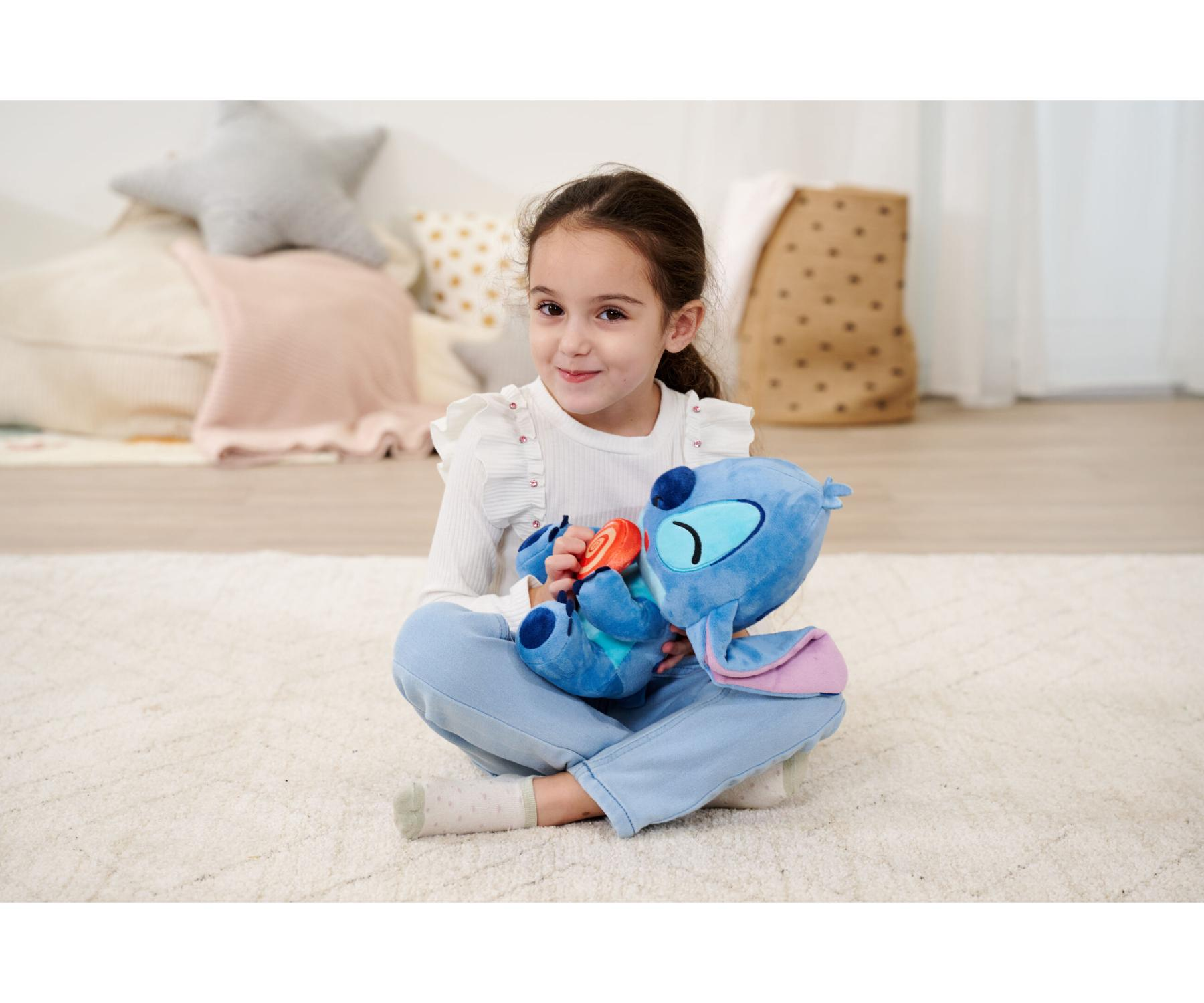 Ein Mädchen sitzt auf einem Teppich und hält ein Stitch-Plüschtier und ein Bonbon. Der Hintergrund hat Kissen und eine Tasche.