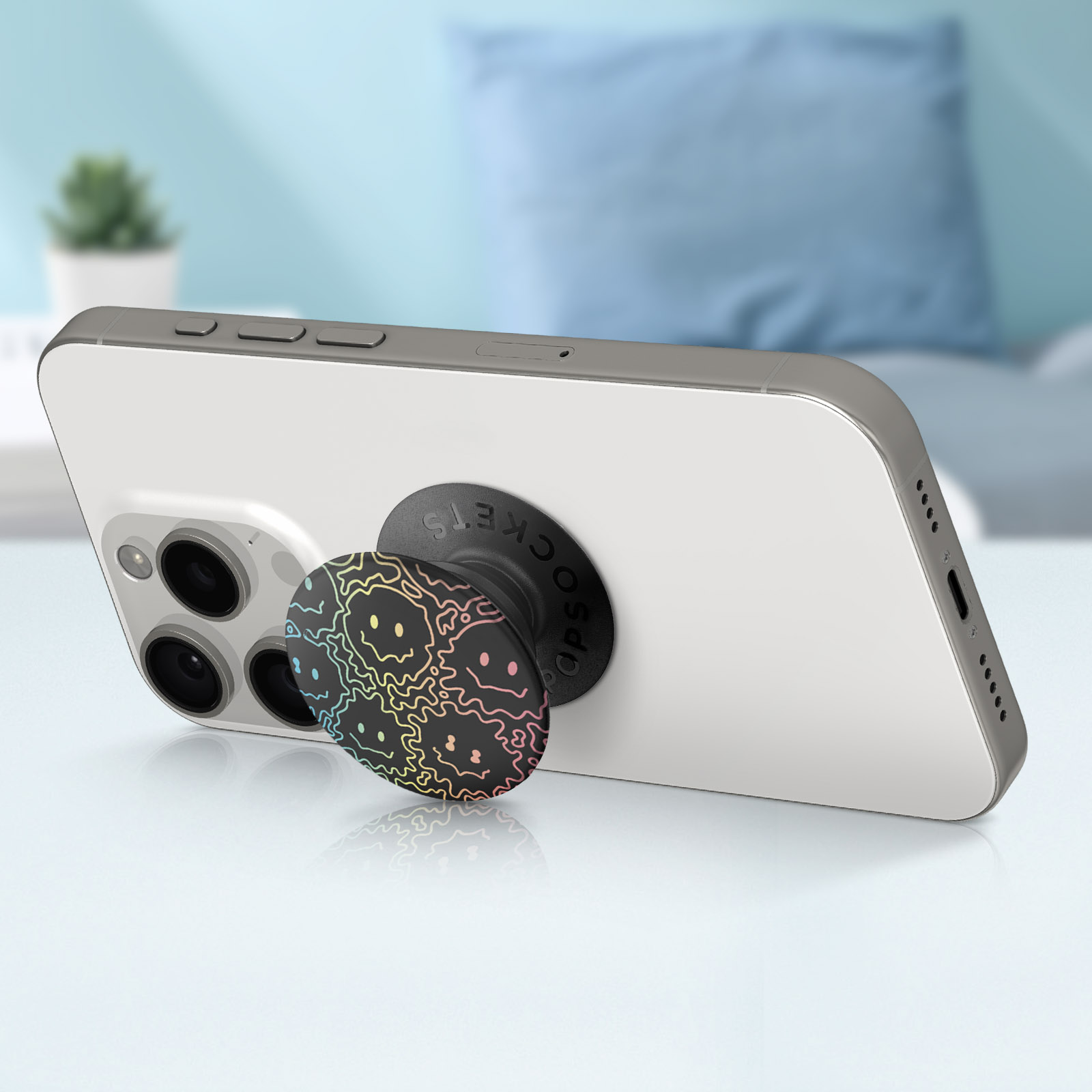 Un iPhone bianco con un PopSocket nero con un design di facce colorate è appoggiato. Lo sfondo è sfocato con un cuscino blu.