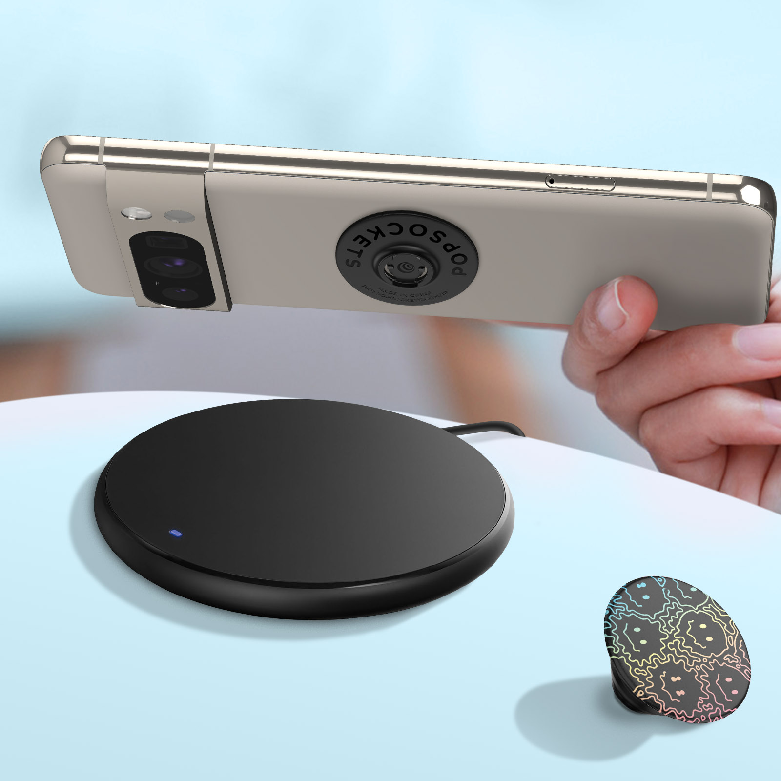 Telefono con PopSockets su un caricatore wireless e un'impugnatura PopSockets staccata. Sfondo azzurro.