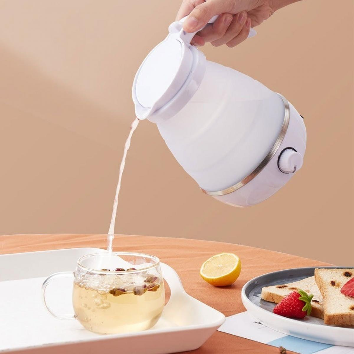 Ein weißer Wasserkocher gießt Wasser in ein Teeglas. Daneben sind ein Teller mit Essen, eine Zitrone und ein Tablett zu sehen.