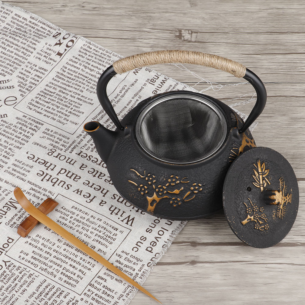 Schwarze Teekanne mit goldenem Blumendesign, Deckel abgenommen. Seilgriff, Netzfilter. Auf Zeitung und Holzoberfläche.