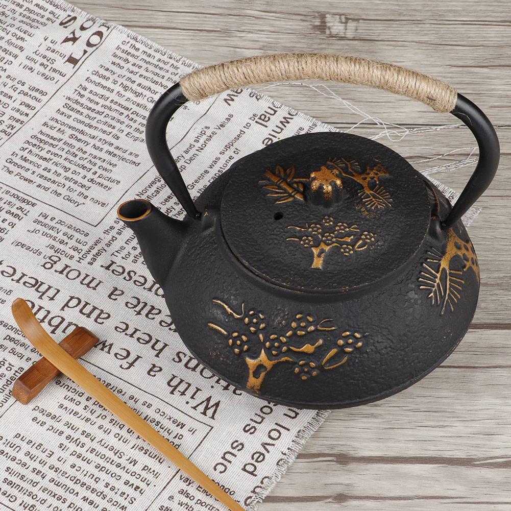 Schwarze gusseiserne Teekanne mit Seilgriff, goldenem Blumendesign, auf einem Zeitungsähnlichen Tuch, mit Holzrührer.