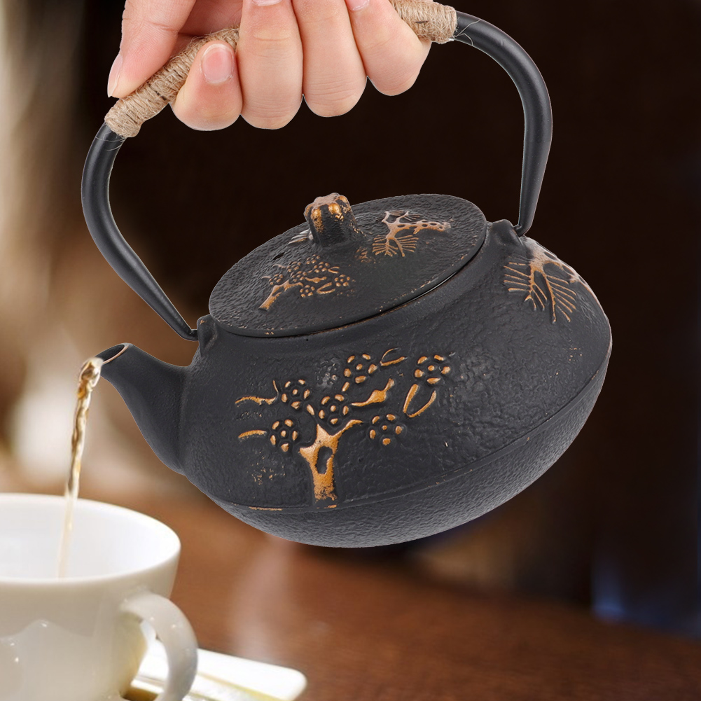 Eine Hand hält eine schwarze Teekanne und gießt Tee in eine weiße Tasse. Die Teekanne hat einen seilumwickelten Griff und ein goldenes Baumdesign.