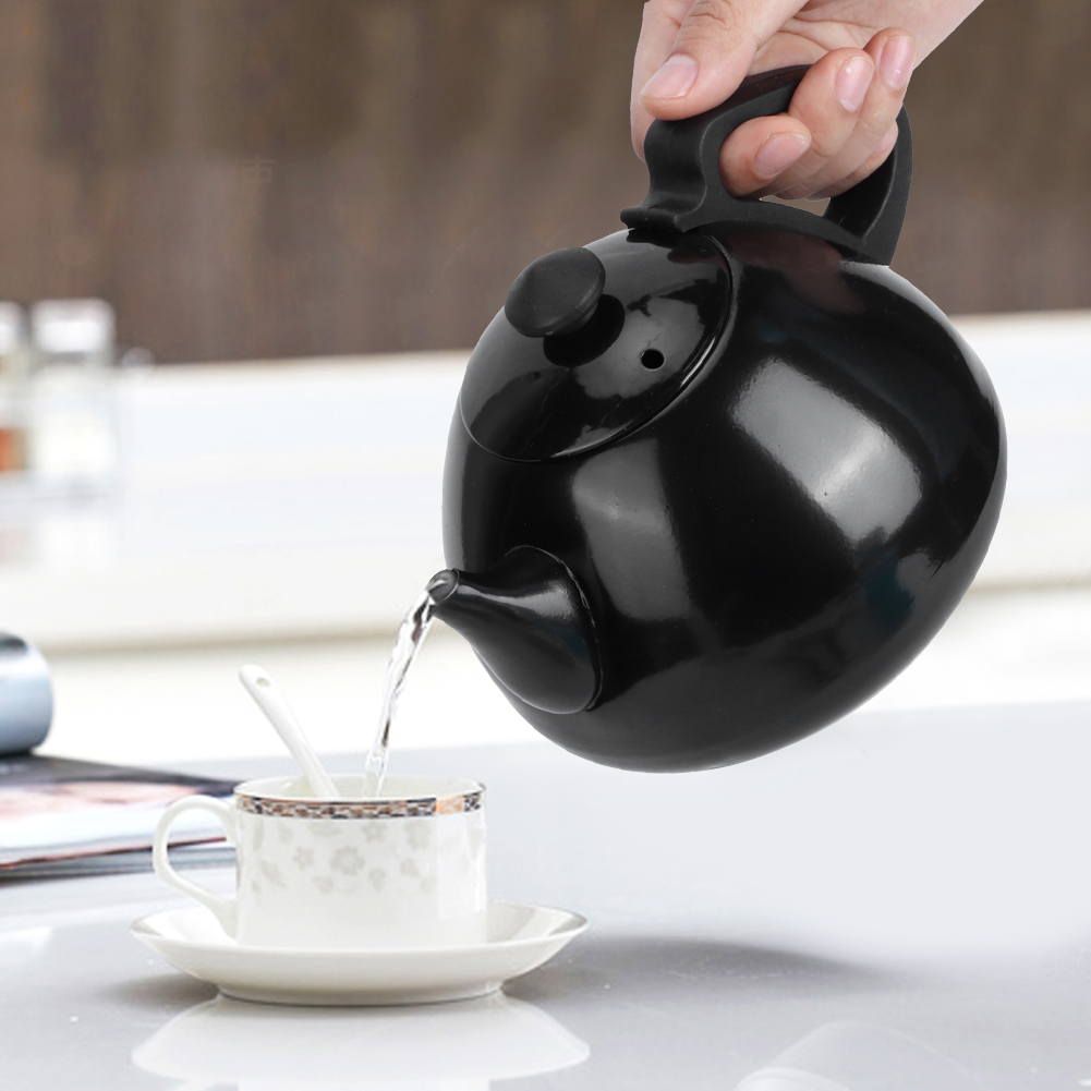 Eine Hand gießt Wasser aus einer schwarzen Teekanne in eine Tasse mit Untertasse. Die Tasse hat ein Blumendesign, und ein Löffel liegt darin.