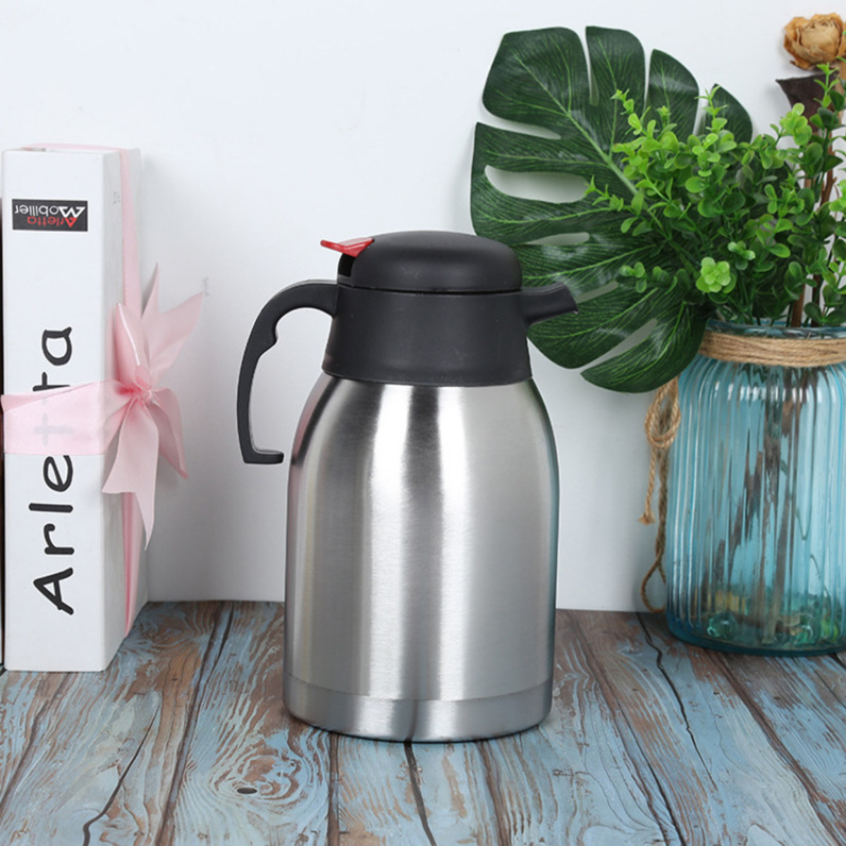 Eine Thermoskanne aus Stahl mit schwarzem Deckel und Griff. Daneben ein Buch mit rosa Schleife und eine Vase mit Pflanzen.