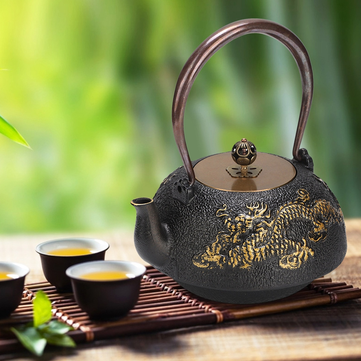 Eine schwarze Teekanne mit Drachendesign und goldenen Akzenten steht auf einer Bambusmatte mit Teetassen. Grüner Bokeh-Hintergrund.