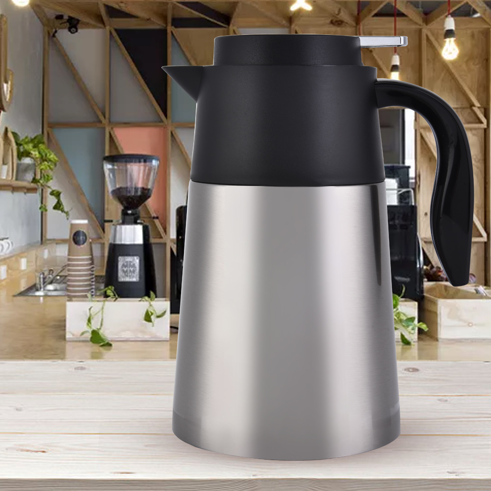 Eine metallische Kaffeekanne mit schwarzem Deckel und Griff steht auf einem Holztisch. Eine Kaffeemühle und Tassen sind im Hintergrund.