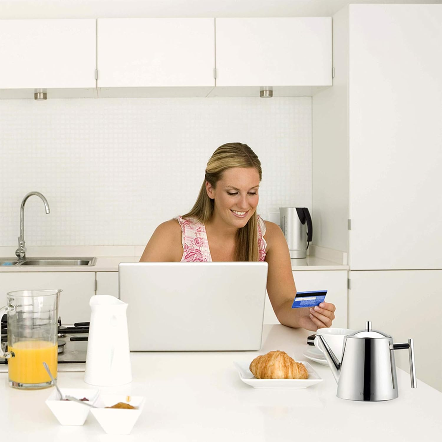 Frau in der Küche mit Laptop und Kreditkarte. Frühstücksartikel auf dem Tisch. Weiße Schränke und Geräte sind sichtbar.