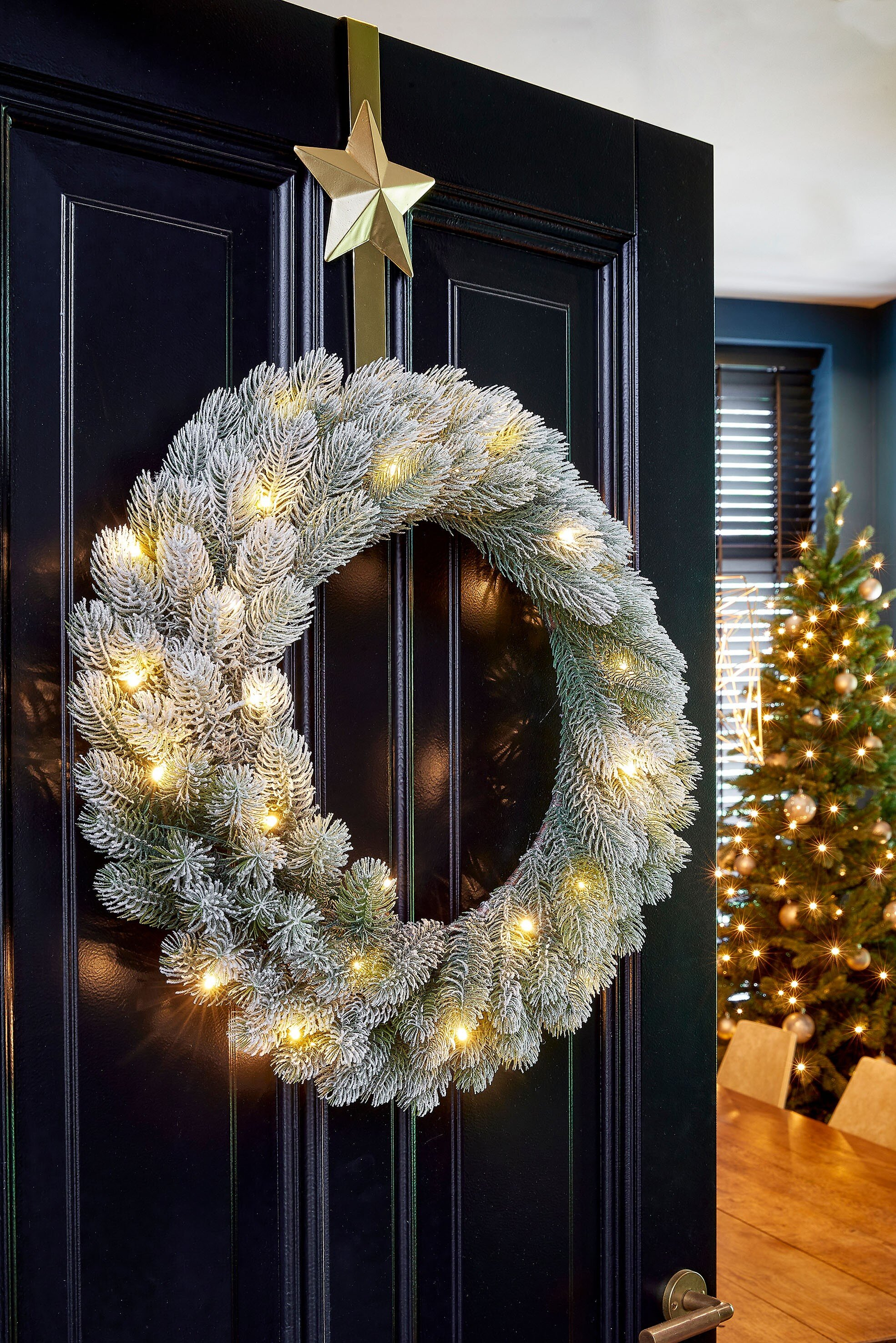 Une couronne de Noël avec des lumières est suspendue à une porte noire, avec un arbre et une table en arrière-plan.