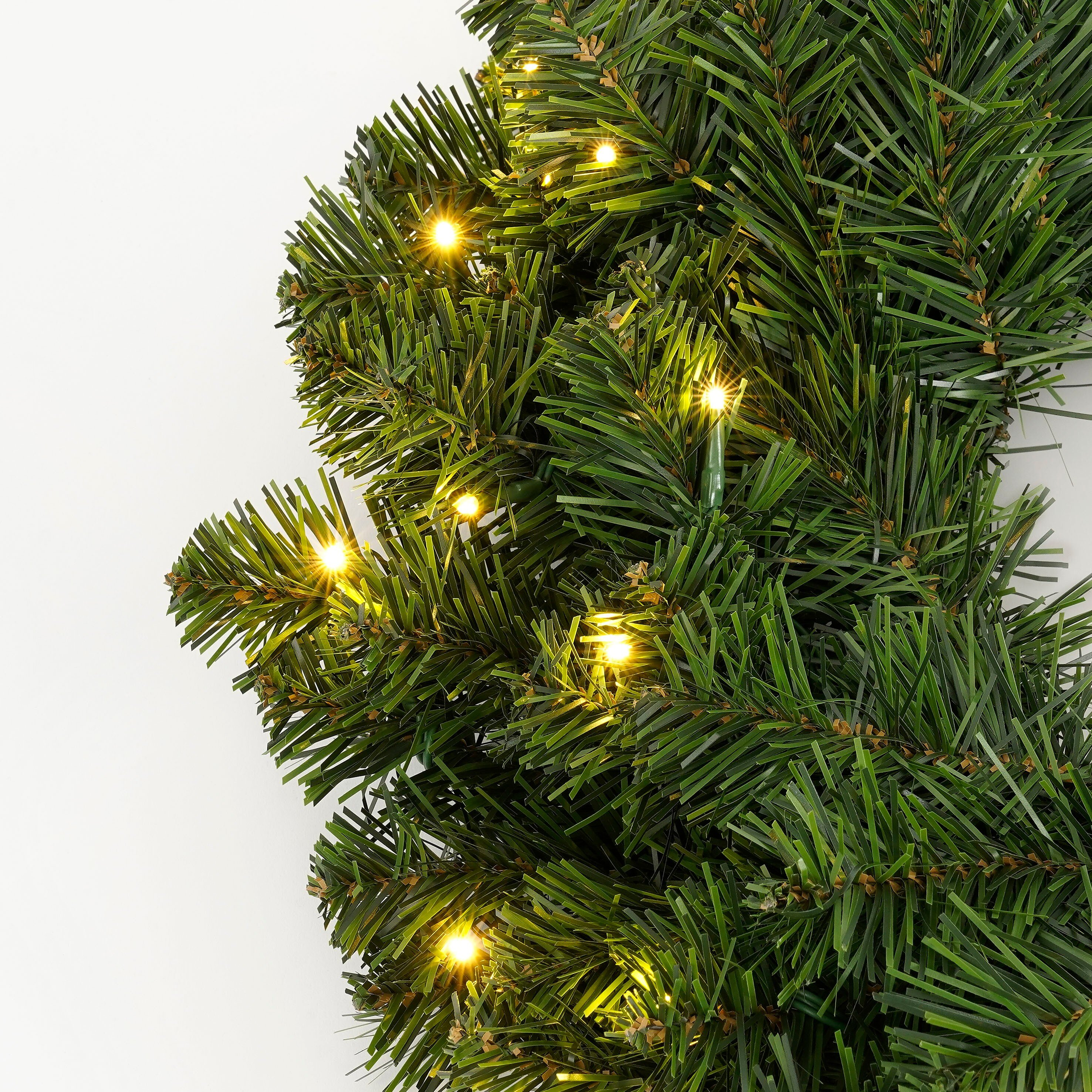 Gros plan d'une guirlande de Noël avec des aiguilles vertes et de petites lumières jaunes, sur fond blanc.