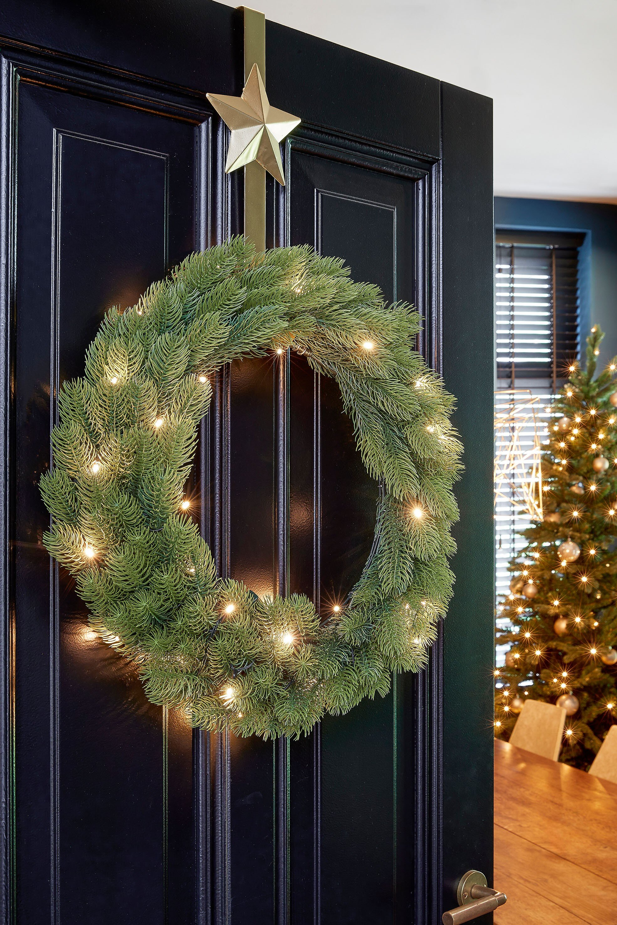 Une couronne de Noël avec des lumières est accrochée à une porte noire. Une étoile dorée décore le haut. Un sapin de Noël en arrière-plan.