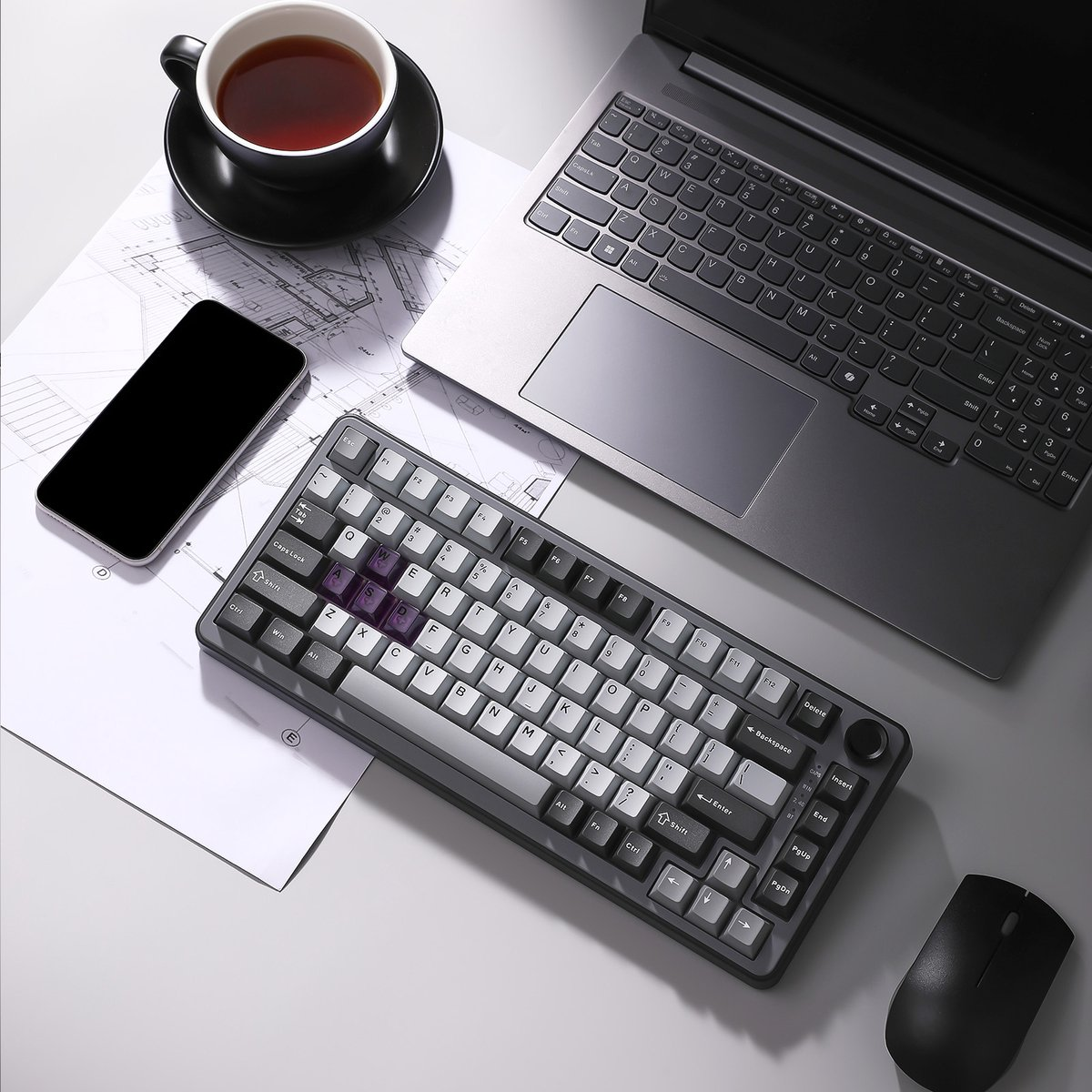 Een bureau-opstelling met een laptop, toetsenbord, muis, telefoon en een kopje thee. Toetsen grijs en wit.