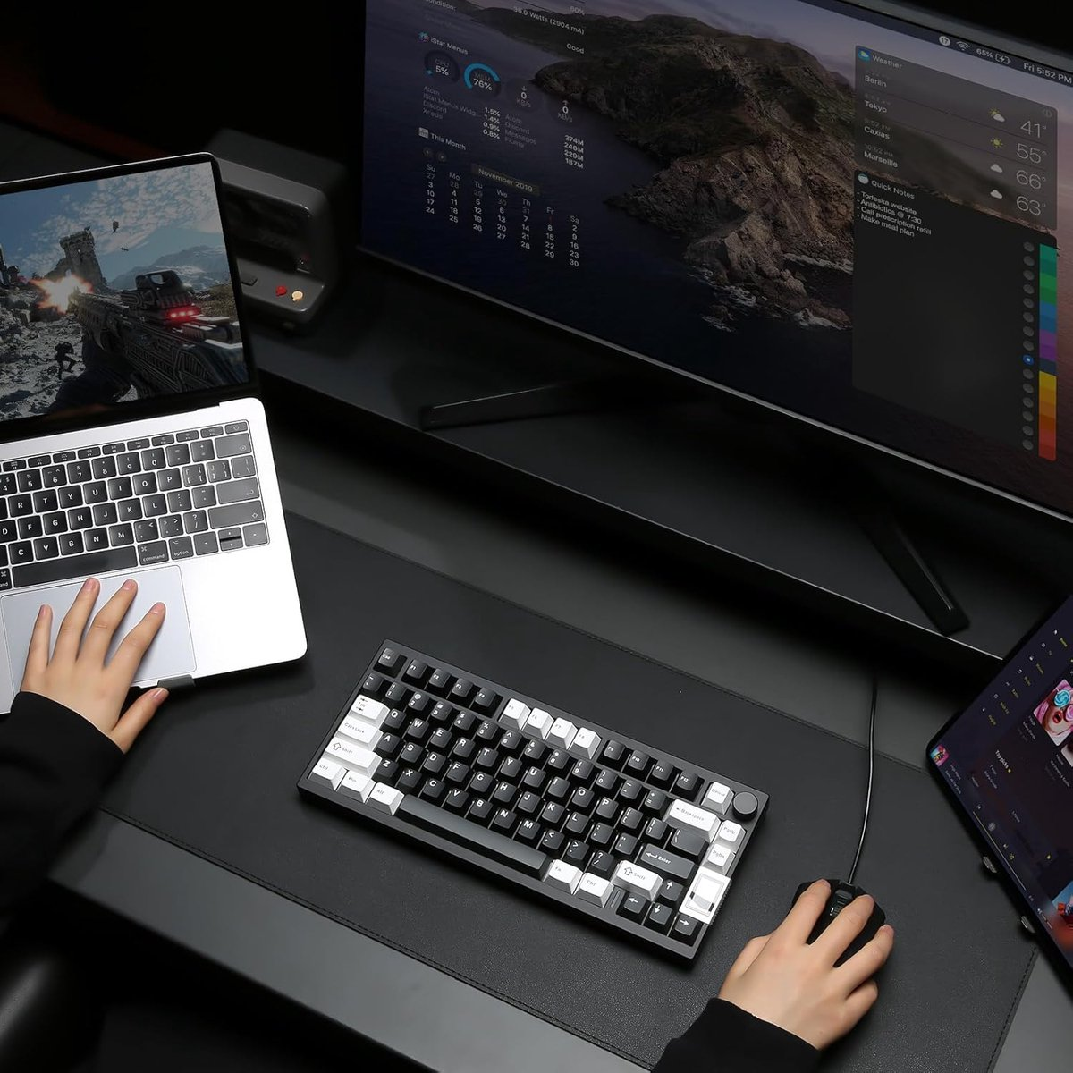 A person uses a laptop, keyboard, and mouse on a desk with a monitor and a tablet. The scene is dark.