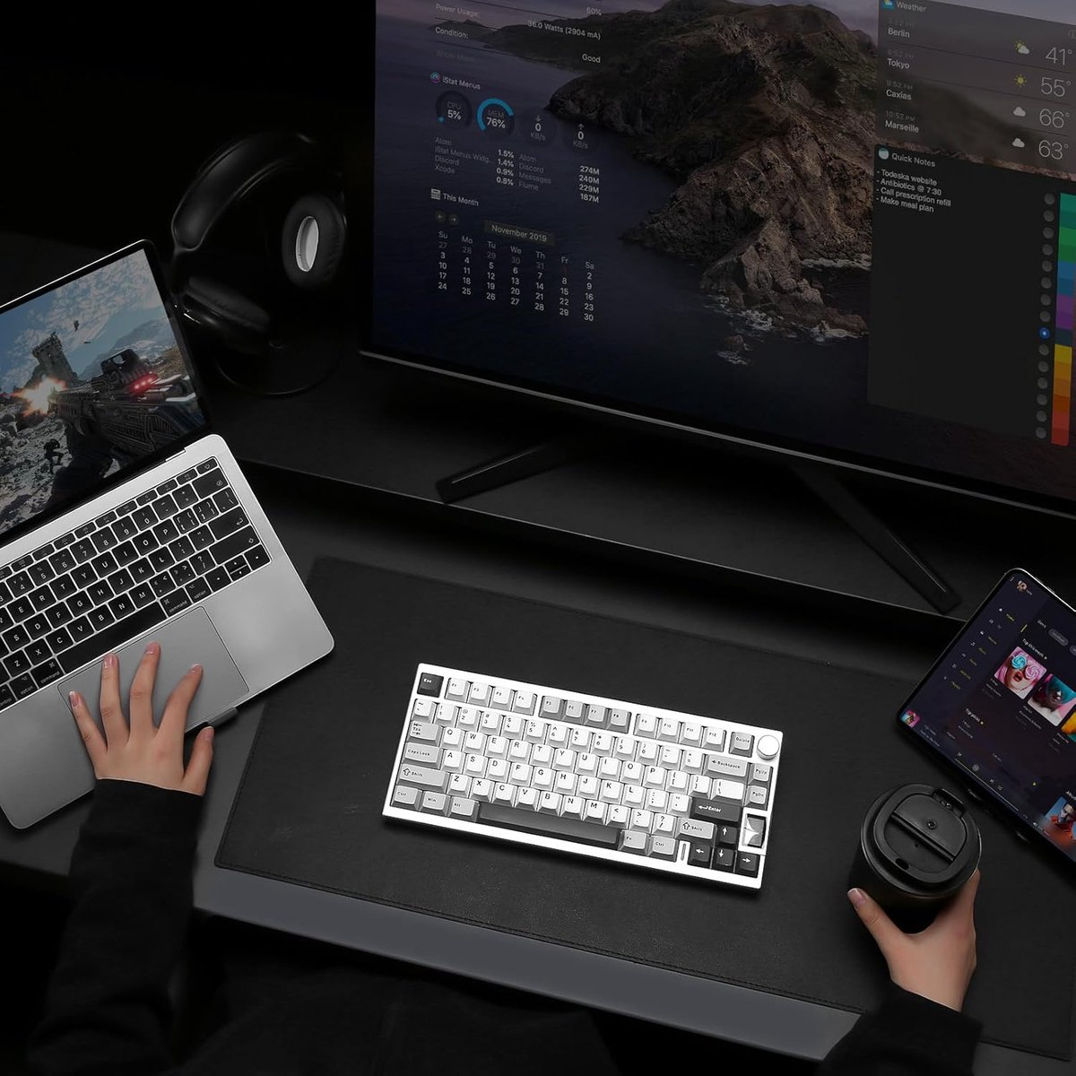 A desk with a laptop, keyboard, tablet, headphones, and a coffee cup. A person's hands are visible.