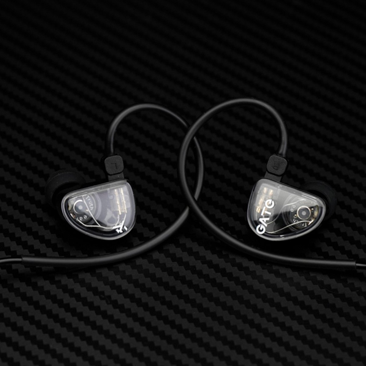 Twee zwarte in-ear monitors op een zwart oppervlak met textuur. Elk heeft een doorzichtige oortelefoon met zichtbare interne componenten en een zwarte kabel.