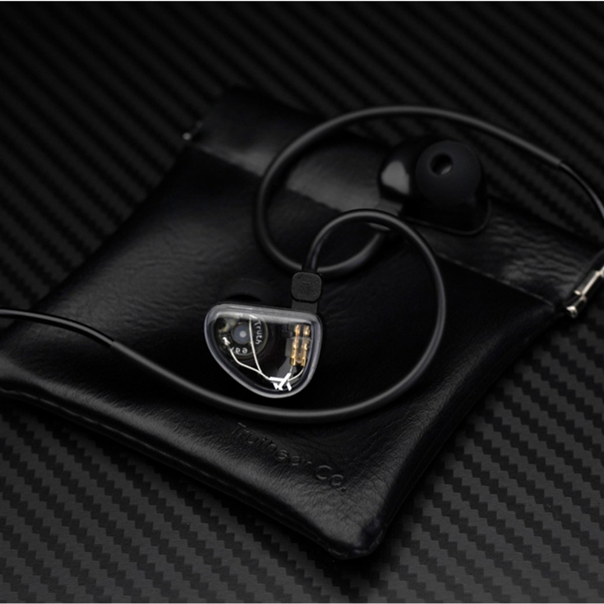 Zwarte in-ear hoofdtelefoon op een zwarte leren etui, geplaatst op een koolstofvezel oppervlak. De hoofdtelefoon heeft een transparante behuizing.