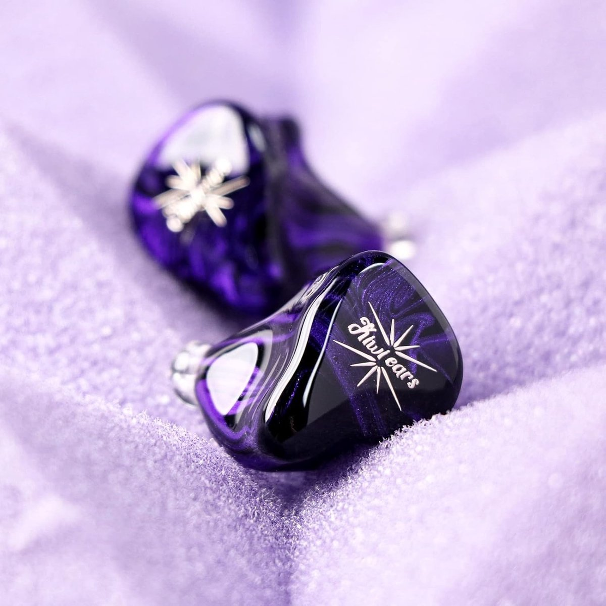 Deux écouteurs Kiwi Ears violets et noirs sur une surface violette texturée. Les écouteurs ont un motif d'étoile et le nom de la marque.