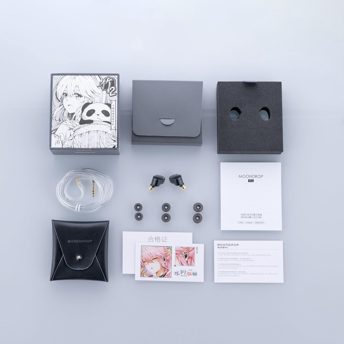 Een bovenaanzicht toont een Moondrop oortelefoonset. Het bevat een doos met anime-kunst, oortelefoons en accessoires. De set staat op een grijs oppervlak.