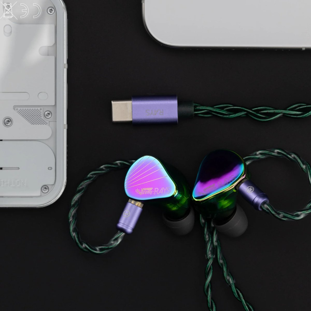 Oordopjes, een USB-C-kabel en een gedeeltelijk zichtbaar apparaat liggen op een zwart oppervlak. De oordopjes zijn iriserend, de kabel is paars.