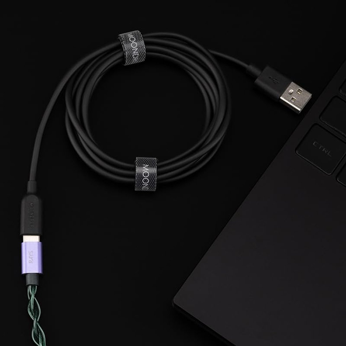 Een zwarte kabel, opgerold, met twee Moon-banden. Een USB-connector en een paars gekleurde connector zijn zichtbaar, samen met een laptop.
